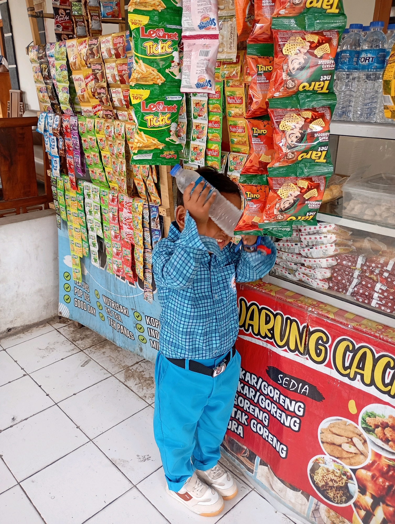 A young boy, wearing a blue checkered shirt and blue pants, stands in front of a street food stall in Babelan, Indonesia. He is holding a partially full plastic water bottle to his forehead, appearing to be cooling himself. The stall displays a variety of snacks and bottled drinks on shelves. The boy's shoes are white with beige accents and the inscription "Sport". The stall itself is decorated with a red banner that advertises "SEDIA KAR/GORENG" (Indonesian for "Available Fried/Fried Snacks") and features images of fried foods. The ground is tiled.