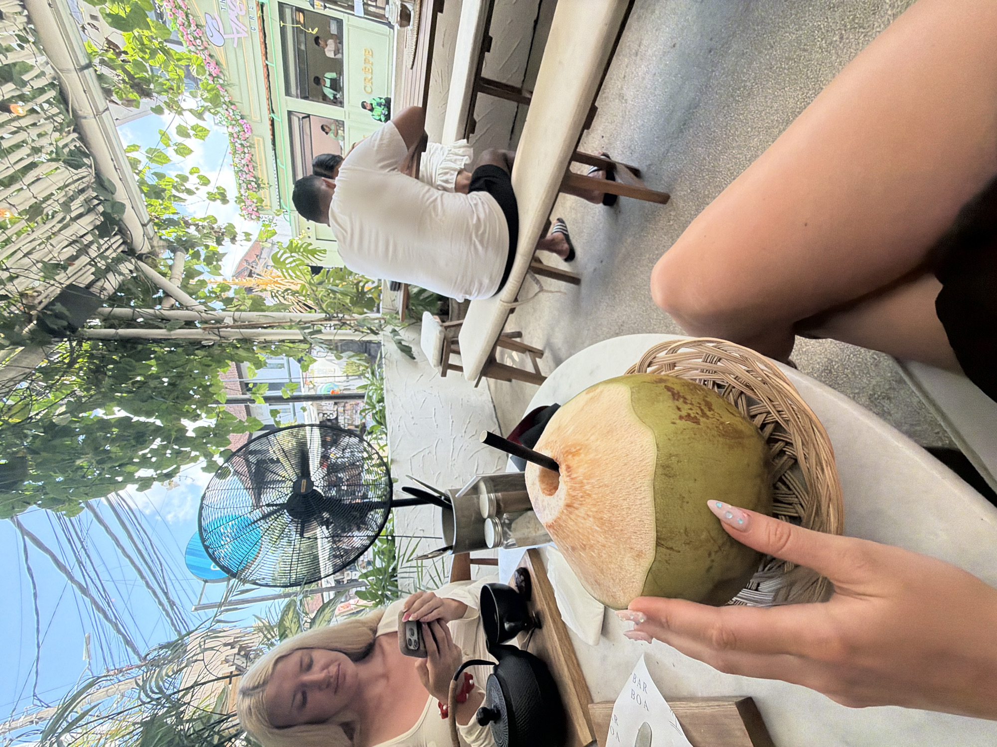 A close-up, POV shot captures a table at an outdoor cafe in North Kuta, Indonesia, on a bright, sunny day. The foreground is dominated by a whole coconut with a straw, resting on a woven placemat. To the left, a woman with blonde hair, wearing a white top, looks down at her phone. A black teapot and a black cup sit on a wooden board beside her. In the midground, a man in a white t-shirt and black shorts is seated on a long bench, facing away from the camera. Behind him, a green food truck or cart with "CREPE" visible on its side is parked. The background features lush green foliage, a large industrial fan, and overhead wires against a clear blue sky, suggesting a tropical, urban setting. The overall impression is one of casual relaxation in a vibrant outdoor space.