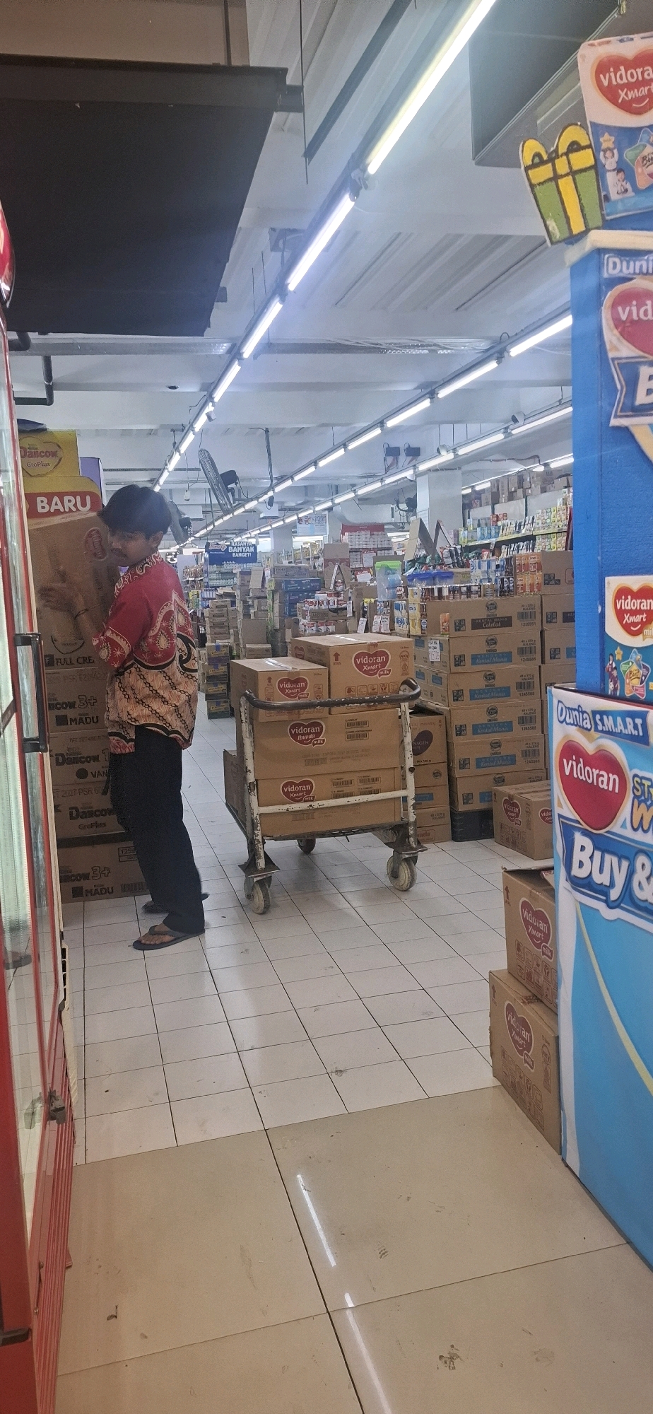 In a retail environment identified as a supermarket in Kartasura, Indonesia, a young male with dark hair wearing a patterned red shirt and dark pants is standing next to a large cardboard box. He is holding onto the box. In the background, there is a shopping cart filled with numerous boxes, some of which are stacked on top of each other. These boxes appear to be merchandise, with labels indicating "vidoran" and "Dancow." The store features fluorescent lighting illuminating the aisles, and shelves stocked with more products are visible in the distance. The floor is tiled in a checkered pattern of white and beige. To the right of the frame, a blue and yellow promotional display stands. The scene suggests a commercial setting, likely a supermarket aisle.