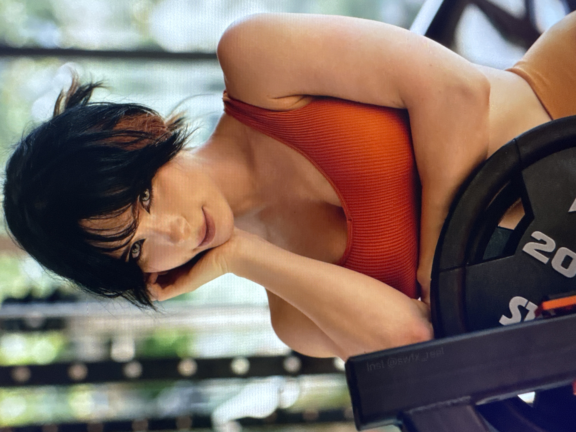 A woman with dark hair, streaked with auburn, lies on her side with her head resting on her hand. She is wearing an orange, ribbed tank top and her gaze is directed at the viewer with a slight smile. In the background, a weight plate with "20" visible is positioned next to her. The scene appears to be indoors, possibly a gym, with blurred green foliage visible through a window. The lighting suggests it is daytime. A black bar with "Inst @svf_real" is visible in the foreground.