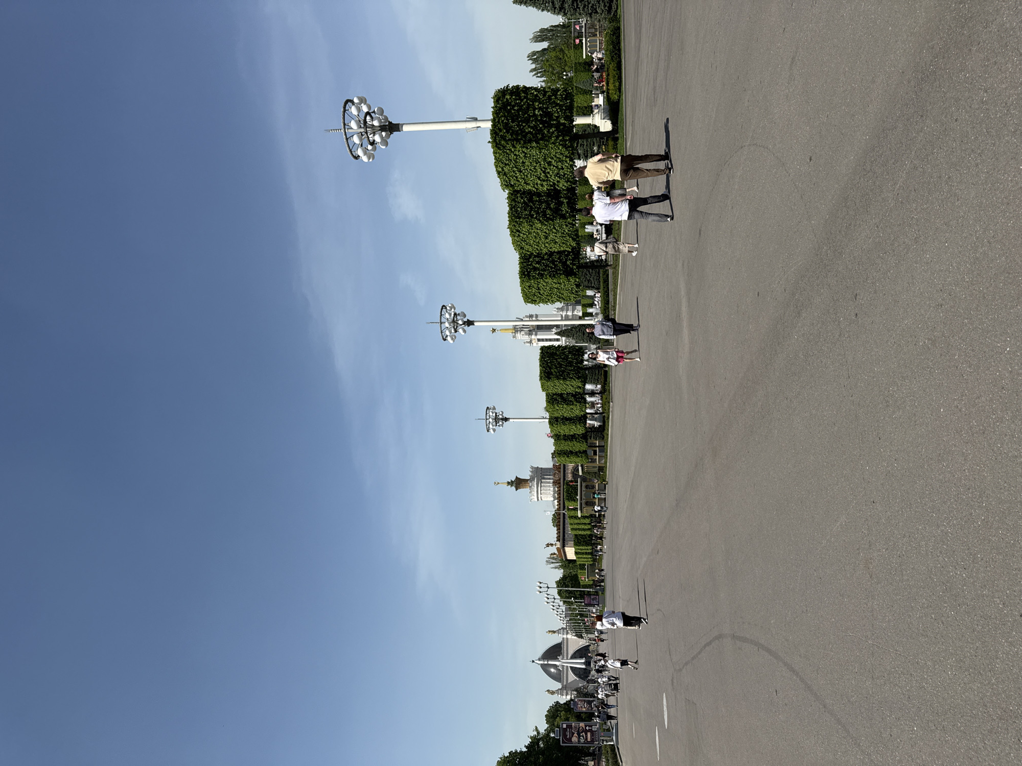 The image shows a long stretch of asphalt, lined with manicured greenery and elaborate streetlights, in Moscow, Russia. Several people are walking along the asphalt. In the background, there are numerous buildings and structures, appearing to be part of a park or exhibition area. The sky is clear and blue, suggesting it's a sunny day. The perspective appears to be from a low angle.