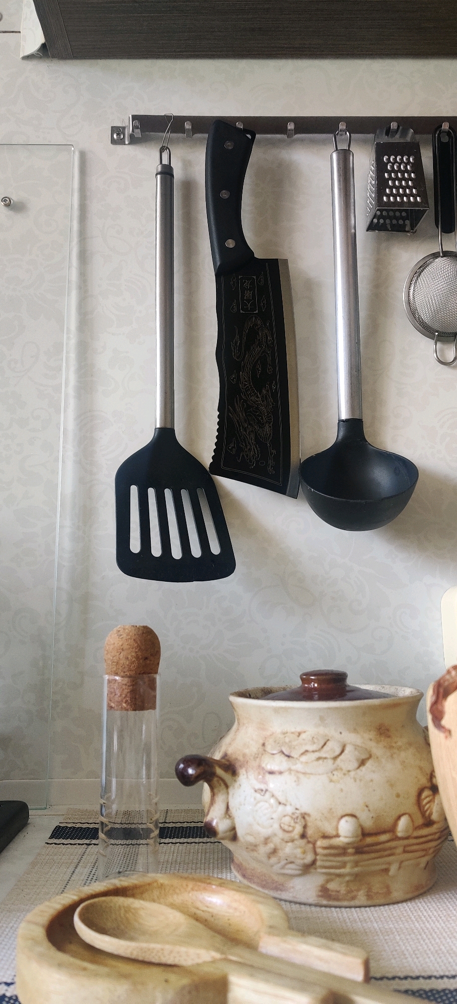 The image depicts a kitchen scene, likely from Hrodna, Belarus, focusing on a collection of kitchen utensils and a rustic pot. Mounted on a wall is a metal rack holding several items. From left to right, these include a black slotted spatula, a large cleaver with a dragon motif etched onto its blade, a ladle, a box grater, and a small mesh sieve. Below the rack, on a striped placemat, sits a tall, slender glass bottle with a cork stopper. To its right is a decorative ceramic pot with handles and an embossed pattern, resembling a scene with figures and fencing. In the foreground, a wooden mortar and pestle holds two wooden spoons. The background is a textured, light-colored wallpaper with a subtle pattern. The lighting suggests an indoor environment, possibly during the day. There are no people or visible actions taking place in the image.