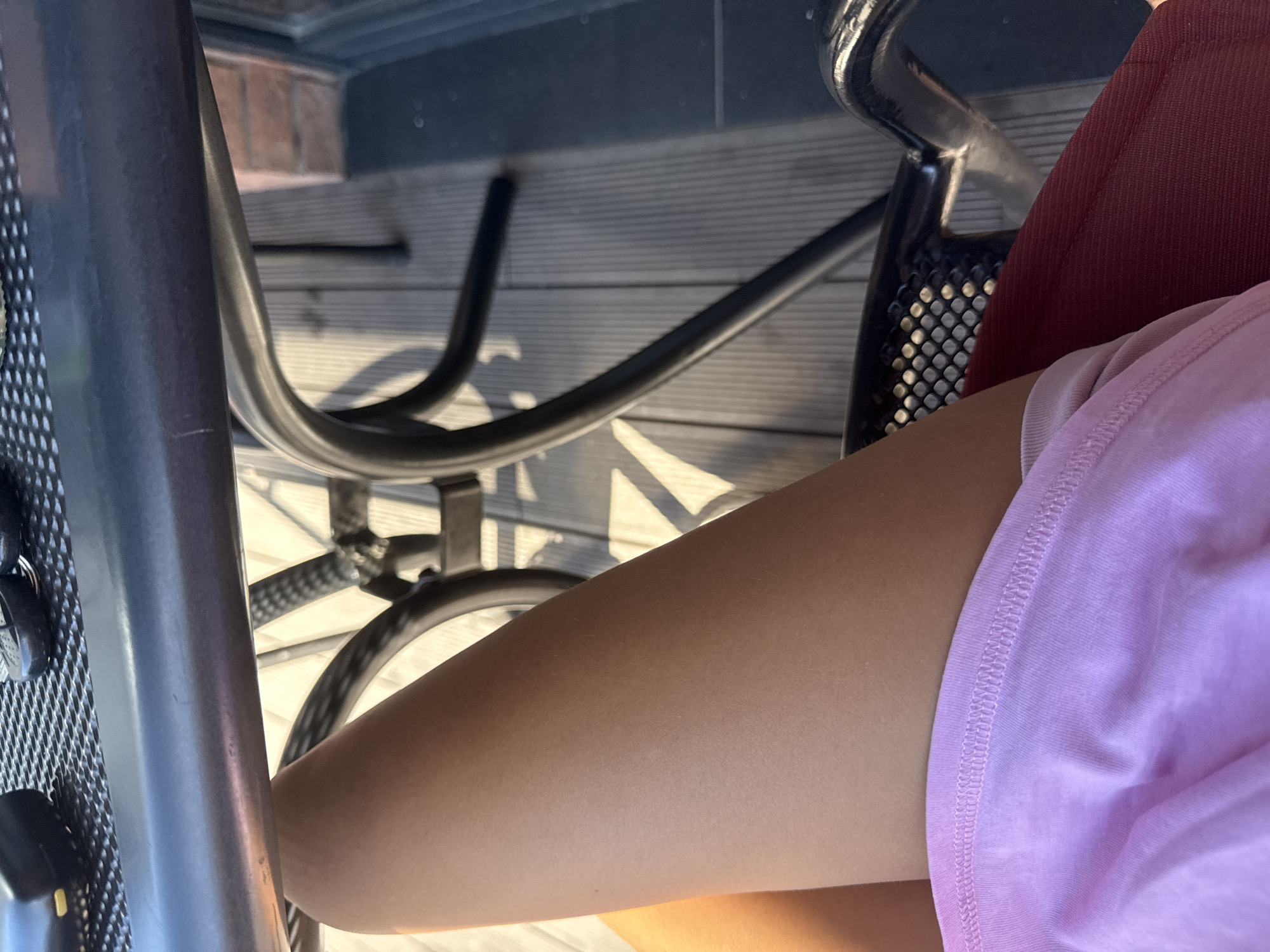 A close-up shot captures a person's legs and thighs, positioned on a chair outdoors. The individual is wearing a light pink t-shirt, and their tanned skin is visible from the knee down. Sunlight casts shadows on a wooden deck, suggesting an outdoor setting. In the background, a metal table with a mesh surface and a black chair are partially visible. The scene appears to be a patio or outdoor seating area, possibly in the city of Comrat, Moldova, given the context of the request.