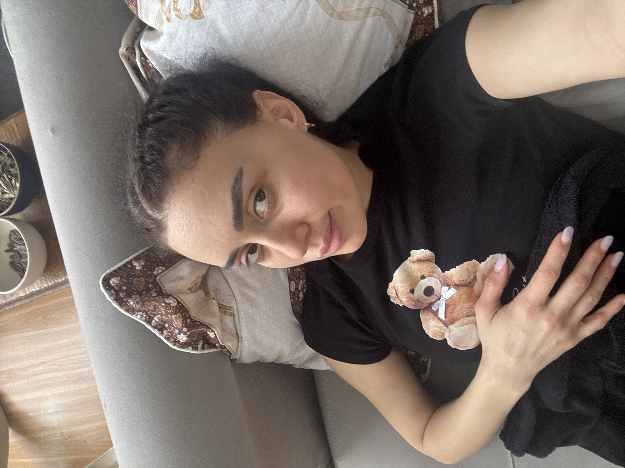 A woman lies on a gray sofa. She is wearing a black t-shirt with a teddy bear graphic. She rests her hand on the bear. A pillow with a patterned border is behind her head. Two pots of plants are visible on a table to the left. The location is not discernible from the image.