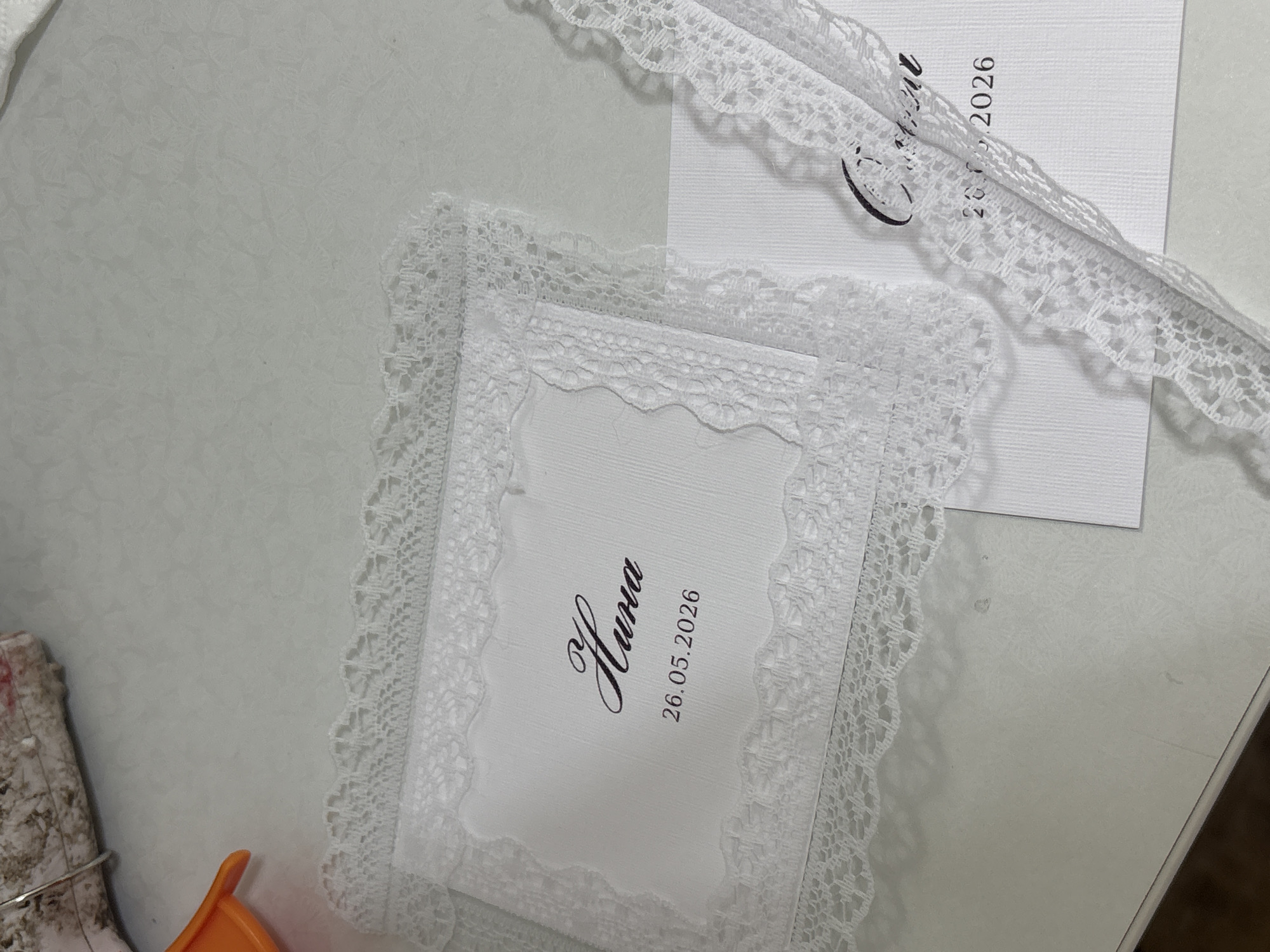 The image shows two white cards with lace borders and an additional length of lace. On the lower left card, the name "Hura" and the date "26.05.2026" are printed in black text. A second card with the date is partly visible at the upper right. The objects are placed on a white surface. No information about the city of Perm, Russia, is present in the image.