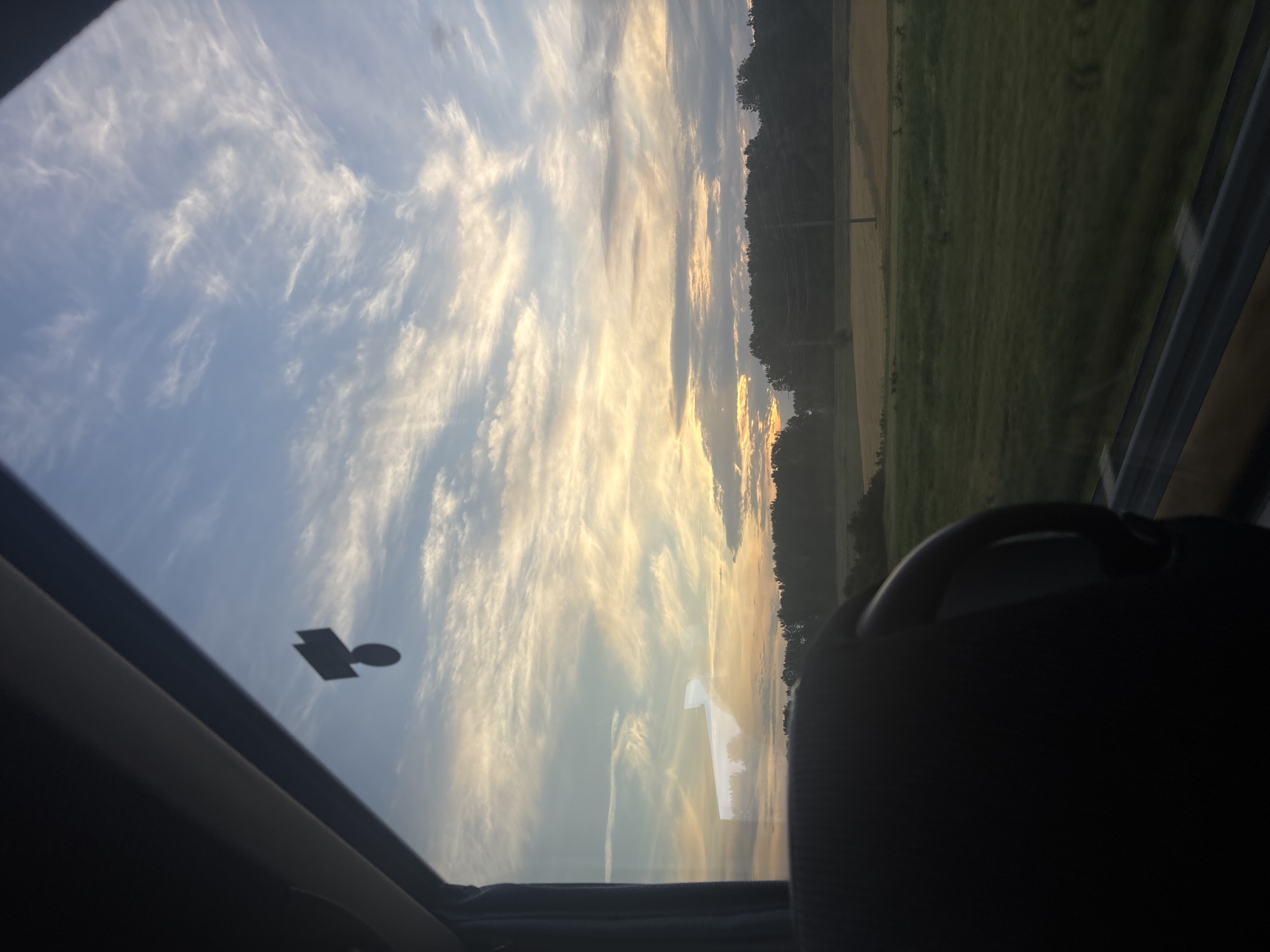 The image shows a view through a car sunroof. The sky is visible, filled with streaky clouds and a golden glow, likely from sunrise or sunset. The edge of a forested area or trees can be seen on the right side, transitioning into a field. A car seat is visible in the foreground, partially obscuring the view. A sticker is stuck to the sunroof. The scene appears to be taken while the car is moving, and shows the road alongside a field or the countryside. I cannot determine the exact location in the city of Obrenovac Urban Municipality, Serbia, from this image.