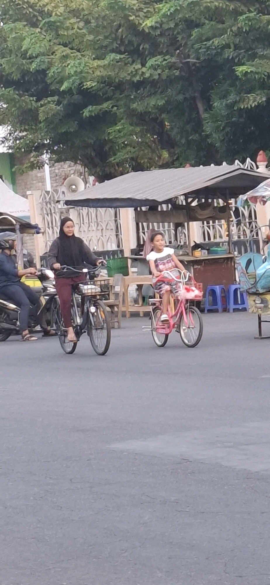 In Desa Donohudan, Indonesia, two individuals are captured in a street scene. On the left, a person wearing a dark hijab and maroon pants rides a bicycle with a basket in front. To their right, a young girl in a colorful dress rides a pink bicycle with a basket. A motorcycle with a rider wearing a helmet is partially visible to the far left. The background features a market stall with a grey roof, some blue stools, and a tree with green foliage. The ground is paved asphalt.
