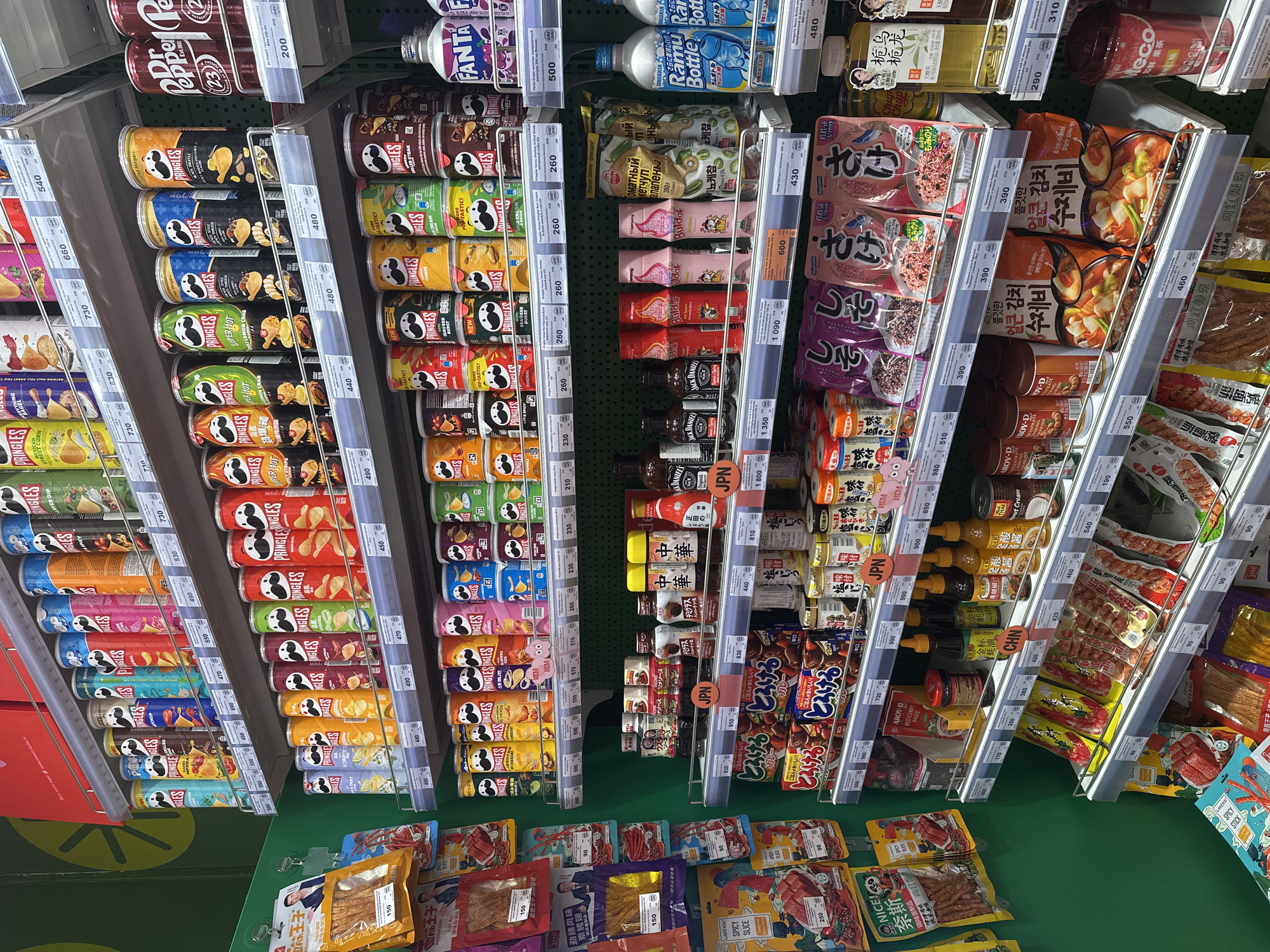 This image captures a high-angle view of a well-stocked supermarket aisle in Moscow, Russia, showcasing a diverse range of international snacks and groceries. No people are visible.
The shelves are dominated by numerous Pringles cans in various flavors, including Paprika, Spicy, Layer Dip, Super Hot, Lemon Crab, Cheese, and Pop-Tastic!. Other prominent beverages include Dr. Pepper, Fanta, Ramune (Ramu Bottle), and Meco fruit tea (如鮮果茶 紅柚).
Beyond the popular brands, a significant section is dedicated to Asian groceries. This includes instant noodles and soups with Korean (쫄깃한 얼큰 김치 수제비) and Japanese (とろけるとろけるカレー, S&B furikake with さけ/salmon and しそ/shiso) text, as well as various sauces and condiments like Jack Daniel's, Aroy-D Red Curry Paste, and Chinese sauces such as "金标黑胡椒汁" (Gold Standard Black Pepper Sauce). Ketchup brands like Buldak and a "Томатный кетчуп алапеньый" (Tomato Ketchup Jalapeno) are also present.
The shelves also display a variety of packaged snacks, notably Asian dried meat/fish products labeled "网红颜条" (internet-famous snacks), "面筋王子" (Mianjin Wangzi), "劲辣风味 素烤翅" (spicy vegetarian grilled wings), and "LAKEXIXI SPICY SLICE".
Products are neatly arranged on metal shelving units with clear price strips indicating prices in Rubles (e.g., 200, 480, 500, 260, 430, 390, 90, 150, 730). Labels indicating country of origin, "JPN" for Japan and "CHN" for China, are visible on some shelves. The bright store lighting highlights the vibrant packaging and extensive product selection, typical of a modern grocery store or an international specialty market.