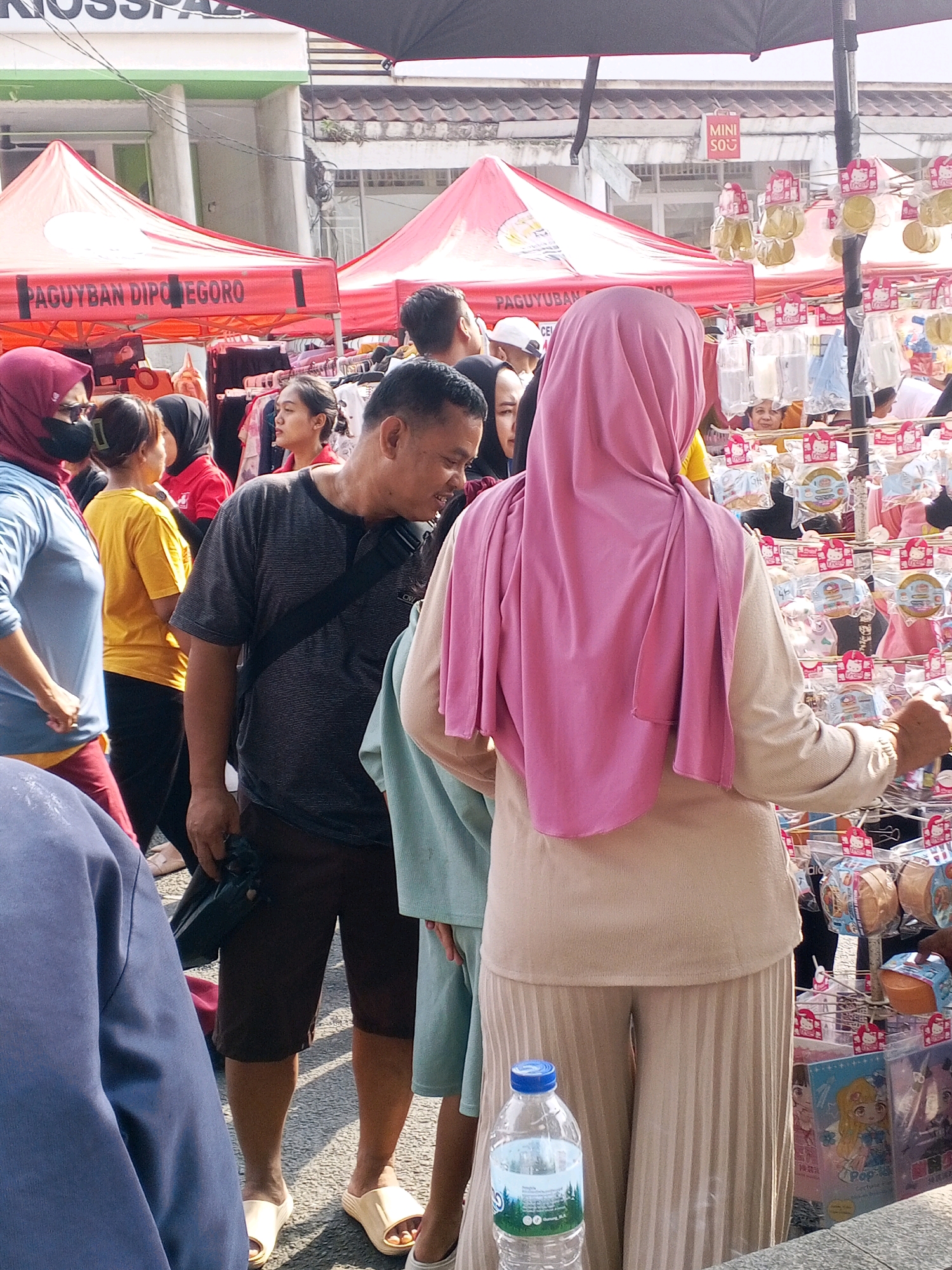 The image depicts a bustling street scene in Serang, Indonesia, likely at a market or fair. In the foreground, a person wearing a light blue hijab and a dark blue jacket is partially visible. To their right, a man in a grey striped t-shirt and brown shorts is looking downwards. Behind him, a woman in a yellow t-shirt and another woman in a black hijab are visible. A large pink hijab worn by a person in a beige outfit dominates the center-right of the frame, with the person's back to the camera. This individual is standing near a stall displaying numerous items packaged in plastic, many adorned with cartoon characters and text in Asian script. These items appear to be small toys or novelty goods. Red tents with the words "PAGUYUBAN DIPONEGORO" printed on them are visible in the background, suggesting a community or organizational event. The ground is paved with asphalt, and overhead, a black umbrella is casting a shadow. The overall impression is one of a vibrant and crowded outdoor gathering.