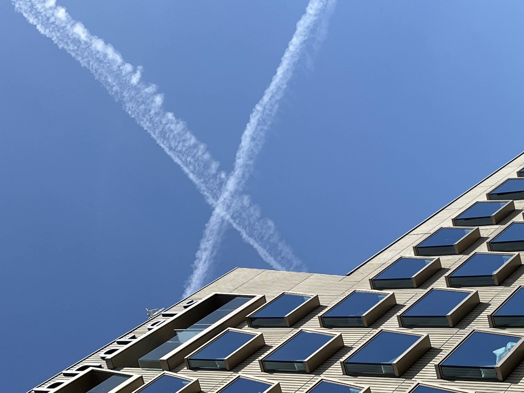 A bright, clear blue sky dominates the upper left portion of the image, where two white, wispy contrails intersect to form an 'X' shape. The contrails are detailed, showing textured, fragmented cloud-like structures. The lower and right sections of the image feature the angular facade of a modern building, observed from a low-angle perspective, looking upwards.
The building's exterior is light beige or tan, with a textured surface composed of horizontal panels. Numerous rectangular windows, or possibly skylights, are set into the sloping facade. These windows feature dark, reflective glass, mirroring the blue sky. They are arranged in a grid-like pattern, staggered across the building's surface as it ascends towards the upper right corner of the frame. The distinct design of the building suggests a contemporary architectural style in Sant Feliu de Llobregat, Spain.
The scene depicts a sunny day with excellent visibility. There are no other clouds present in the sky apart from the contrails. No people or visible text are discernible in the image. The composition highlights the geometric interplay between the natural lines of the contrails and the architectural lines of the building.