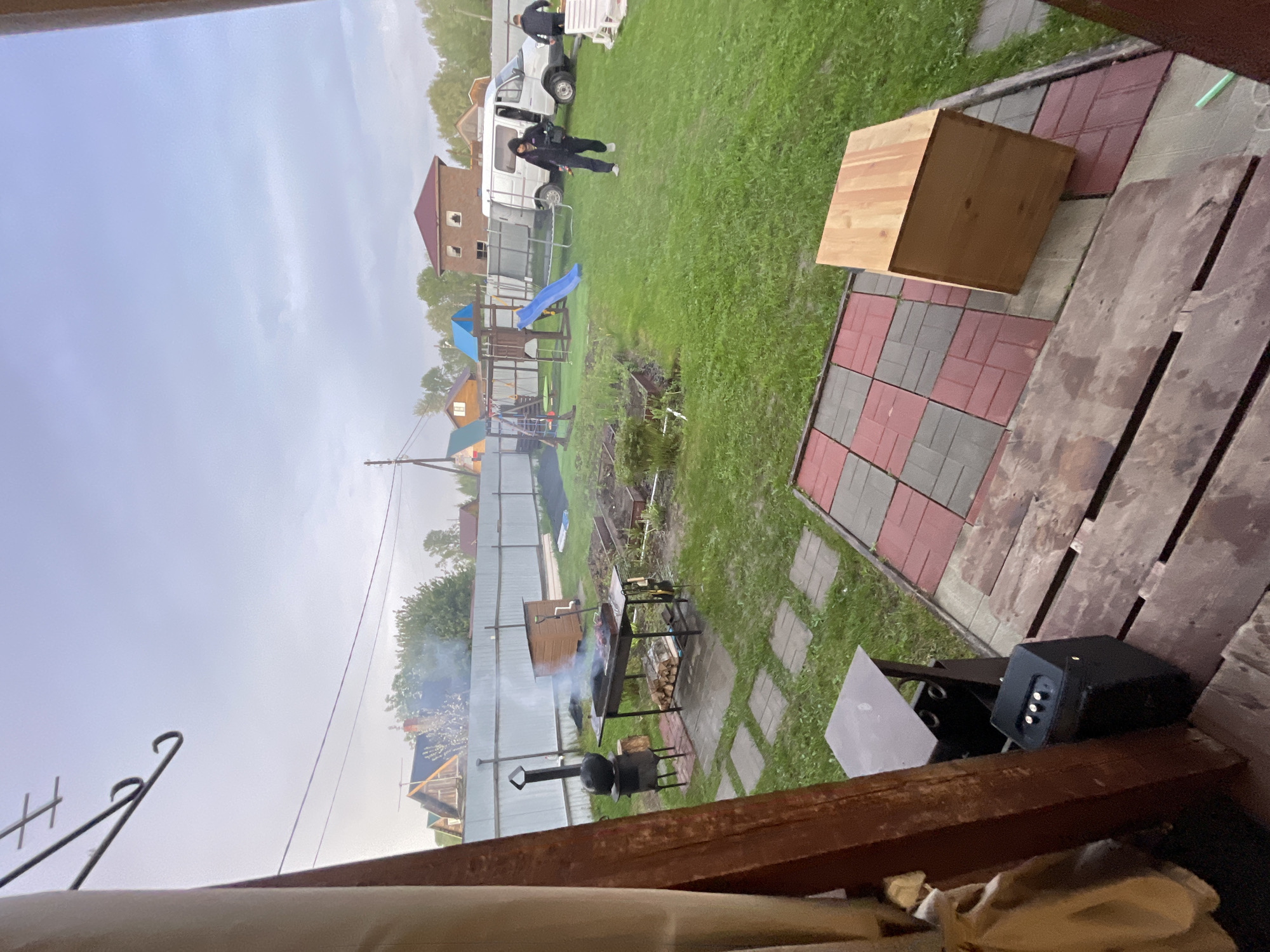 The image depicts a residential scene likely located in a suburban area. In the background, a playground with a slide is visible, adjacent to a white van. Two individuals are standing near the van. A smoker and grill are positioned on a patio area. A wooden box sits on the patio surface. Smoke rises from the smoker and grill, suggesting an active cooking or heating process. The setting appears to be a backyard. It is not possible to definitively confirm the location as Bibikha, Russia.
