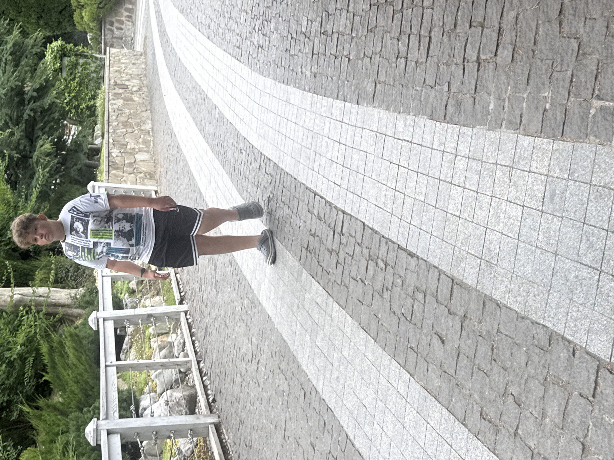 A young boy with curly brown hair walks on a paved pathway. He is wearing a white t-shirt with a collage of images and text, black athletic shorts, and gray slip-on shoes. The pathway is made of grey cobblestones interspersed with white rectangular tiles, forming curved stripes. To the left of the pathway is a white wooden fence with metal wires, behind which are rocks and green foliage. Beyond the fence, there are trees and a stone wall. The background consists of lush greenery and trees. The scene appears to be outdoors in a park or garden area.
