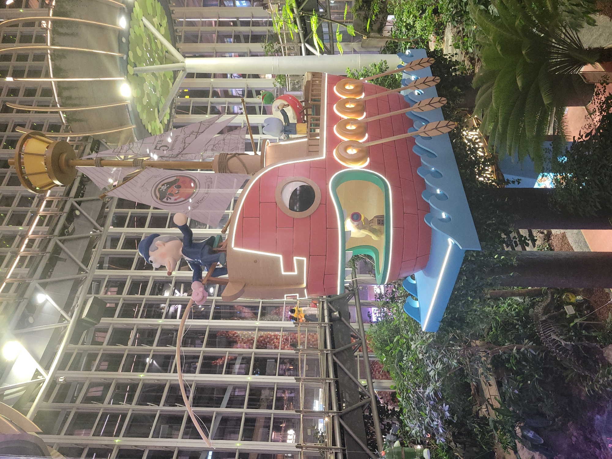 The image shows a large, colorful, cartoon-like pirate ship structure located in an indoor atrium setting. The "ship" features a pink hull, blue "waves," doughnut-shaped decorations, and flags bearing designs. A cartoon figure of a man with a fishing rod sits on a platform near the front of the ship. The ship is situated amongst plants and greenery. Behind the ship, the architecture of the building features a grid of windows. The location of the structure is likely in the Liangjiang district of Chongqing, China based on the context, although this cannot be verified with certainty solely from the image.