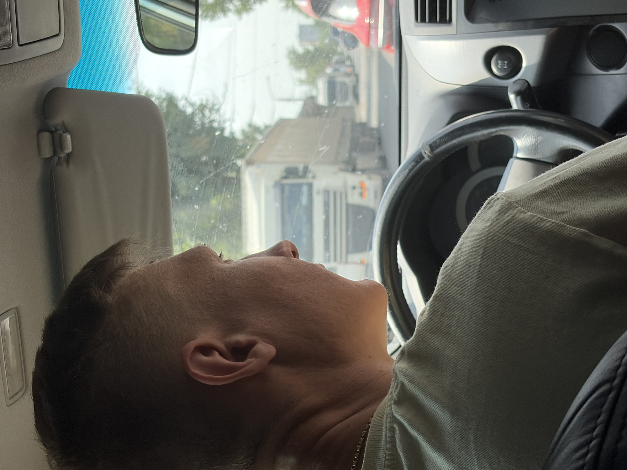 The image shows a man driving a car on a street in Rostov-on-Don, Russia. The man is seen from the side, facing forward, with his left arm resting on the steering wheel. He is wearing a light-colored t-shirt and a gold chain. The interior of the car is visible, including the dashboard, steering wheel, and sun visor. Outside the car, other vehicles are on the road, including a white truck and a red car. The scene appears to be daytime, with sunlight filtering through the trees and buildings in the background.