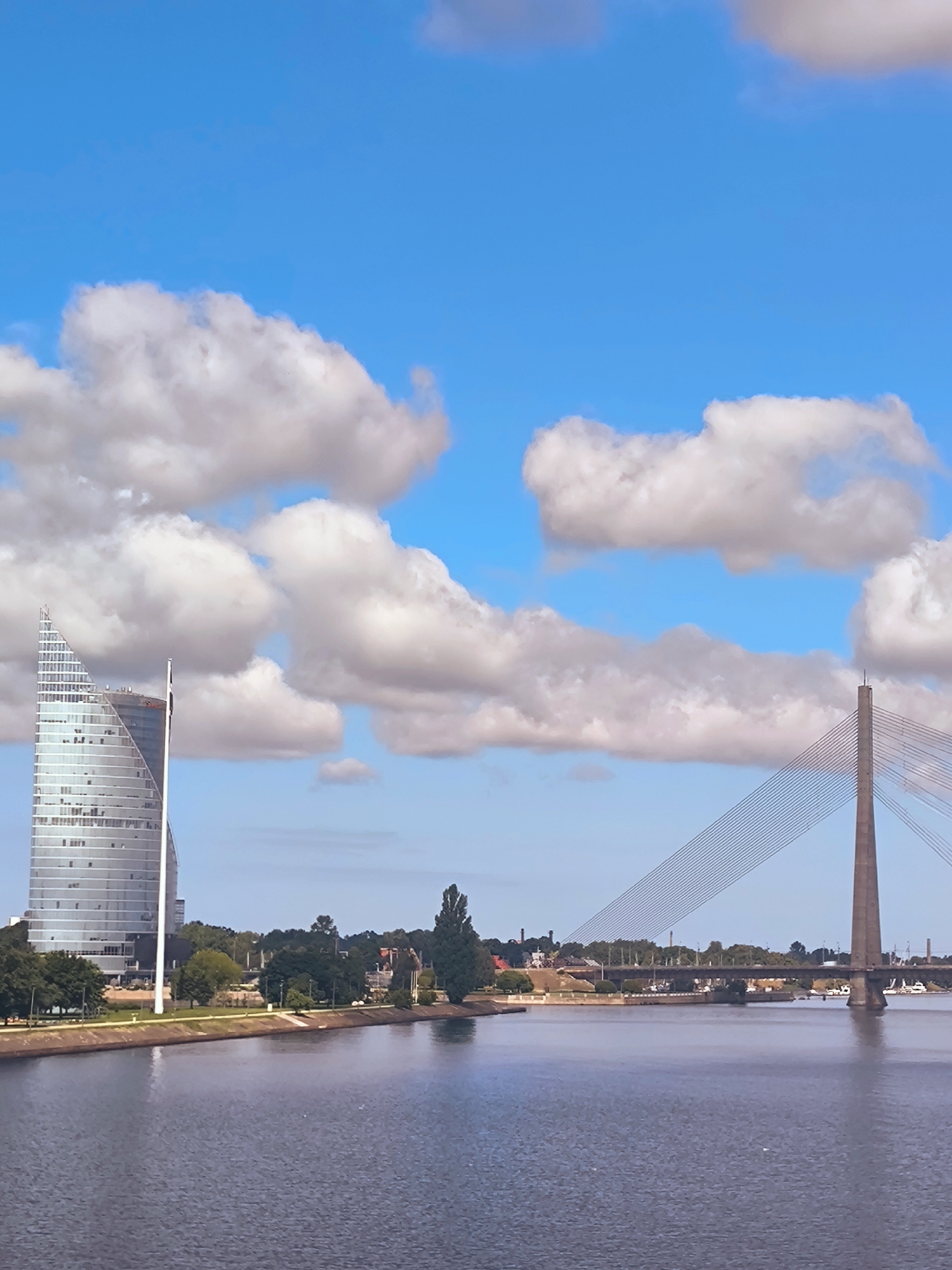 The image depicts a wide river in Riga, Latvia, with a modern skyscraper and a cable-stayed bridge visible on the opposite bank. The sky is blue with fluffy white clouds. The skyscraper, likely the Parex Bank building, has a distinctive triangular shape and a glass facade. The cable-stayed bridge is a prominent landmark, with its tall concrete tower and numerous cables extending across the river. Trees and buildings line the riverbank, suggesting a well-developed urban environment. The water is calm, reflecting the sky and surrounding structures. No people are visible in the scene.