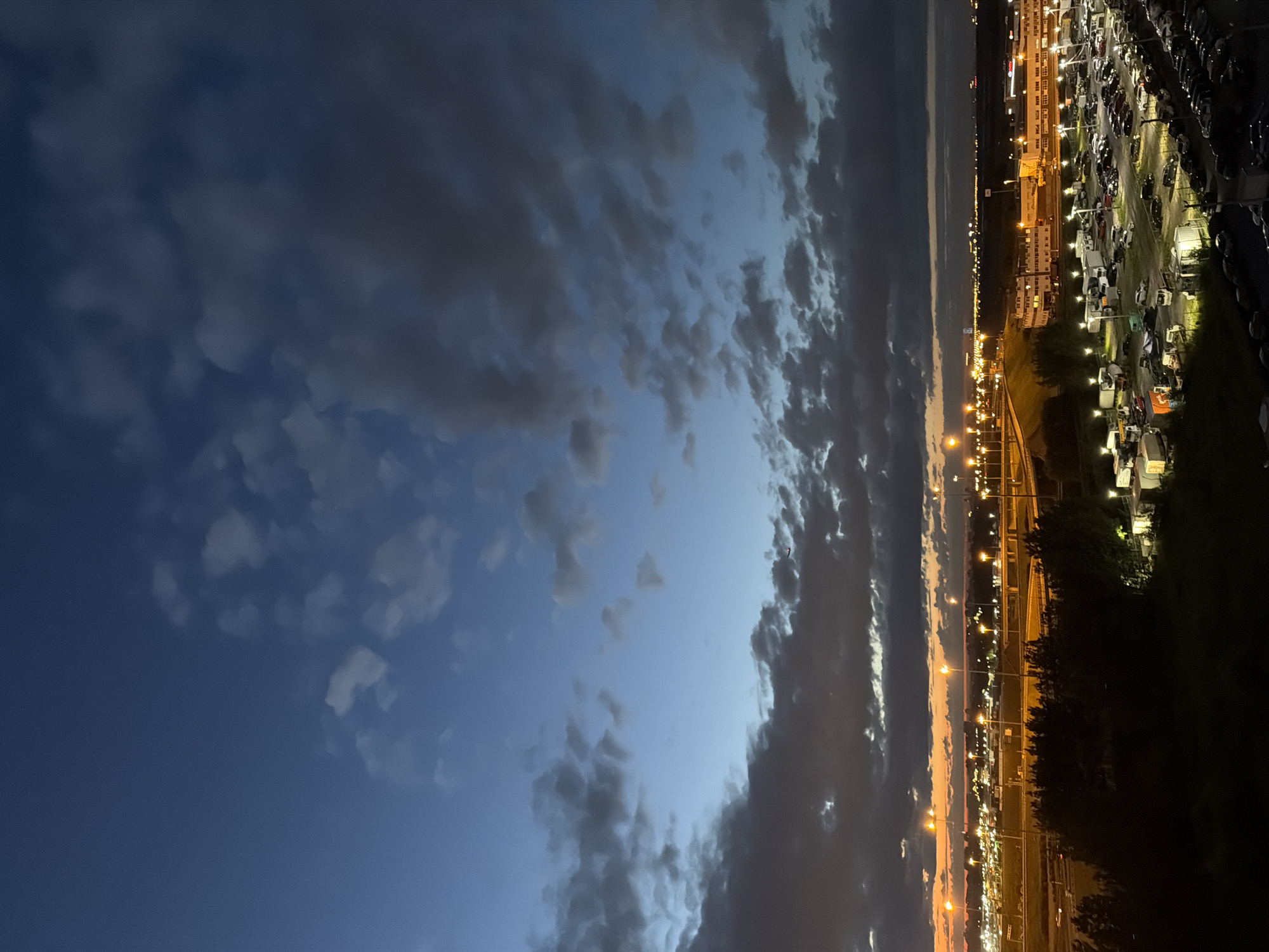 This image captures a twilight scene over an industrial area or airport at dusk. The sky is a deep blue transitioning to lighter shades near the horizon, with scattered clouds. The horizon is marked by a line of bright orange light, suggesting the last rays of the sun. Numerous streetlights and building lights illuminate the landscape below, creating a network of artificial illumination against the darkening sky. In the foreground, there is a large parking lot filled with many cars, and some trucks and trailers are also visible. Trees are present in the mid-ground, partially obscuring the view of some structures. The overall impression is one of a busy, functional area transitioning from day to night, with a dramatic sky overhead.