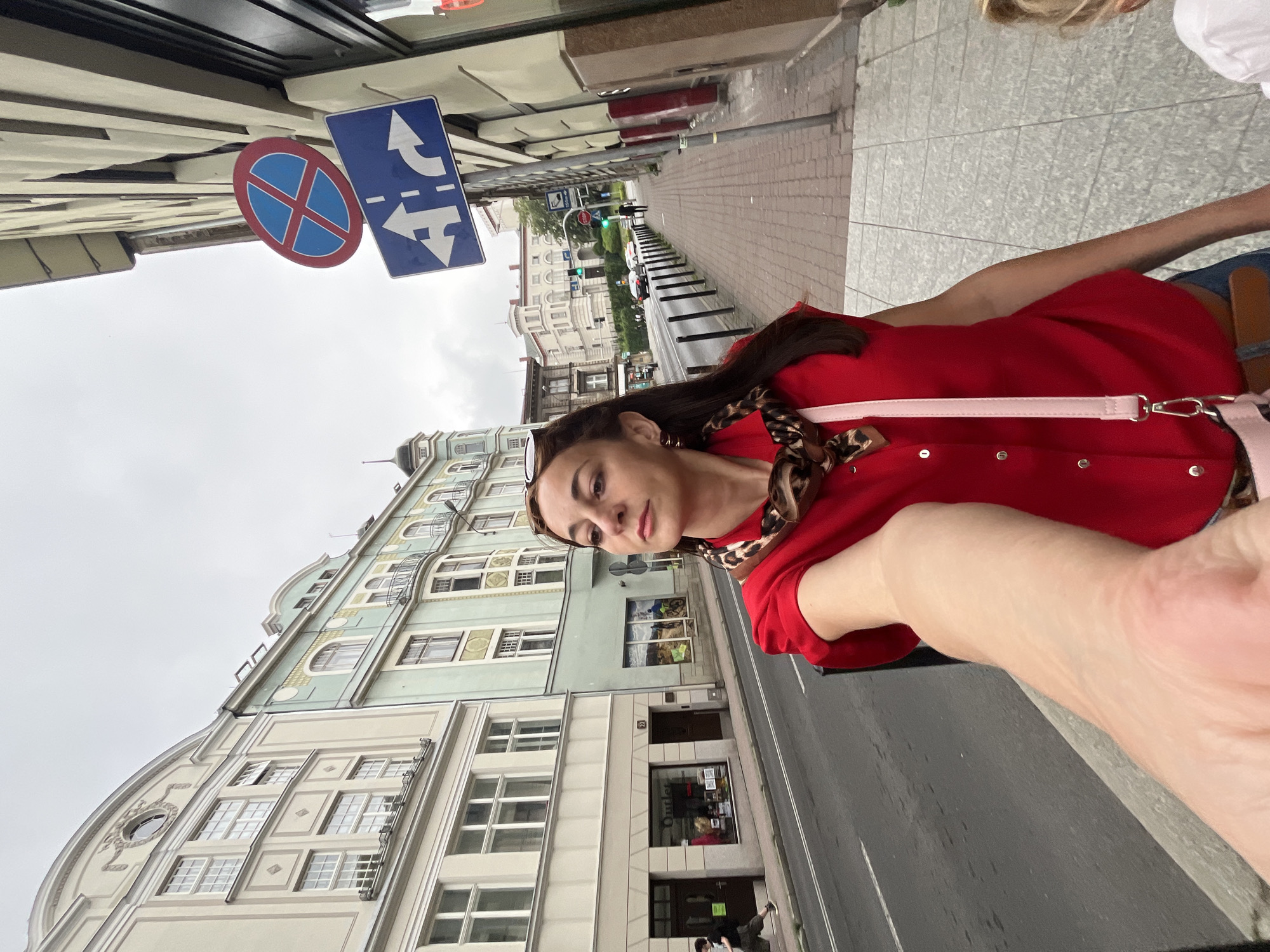 In an outdoor, urban setting, a woman with long brown hair pulled back and a leopard-print scarf around her neck is captured in a selfie. She is wearing a red collared shirt with silver buttons and a light pink purse strap is visible across her chest. Her left arm is extended, holding the camera. A portion of another person's shoulder, with blond hair, is visible in the lower right corner of the frame. The woman stands on a paved sidewalk next to a street with parked cars and passing traffic. Buildings with multiple windows line the street. Two traffic signs are visible on a pole to the right of the woman: one is a red circle with a blue 'X' indicating no stopping, and the other is a blue rectangular sign with white arrows showing traffic lane guidance. The sky is overcast.