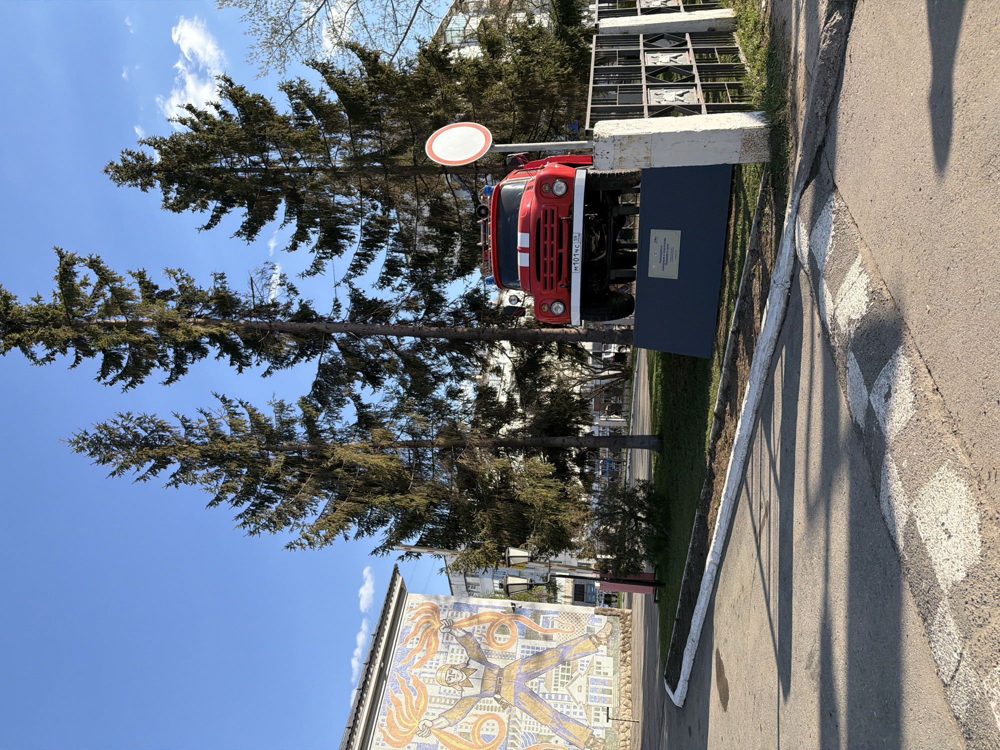 A red fire truck is mounted on a display stand with a metal lattice behind it and a road sign attached, placed in front of two tall pine trees, against a blue sky with scattered clouds. The fire truck has a white stripe across the front, with the license plate visible. A building adorned with a mosaic featuring a human-like figure with arms and legs extended over a cityscape motif is in the lower left corner. The location is the city of Chaikovsky, Russia.