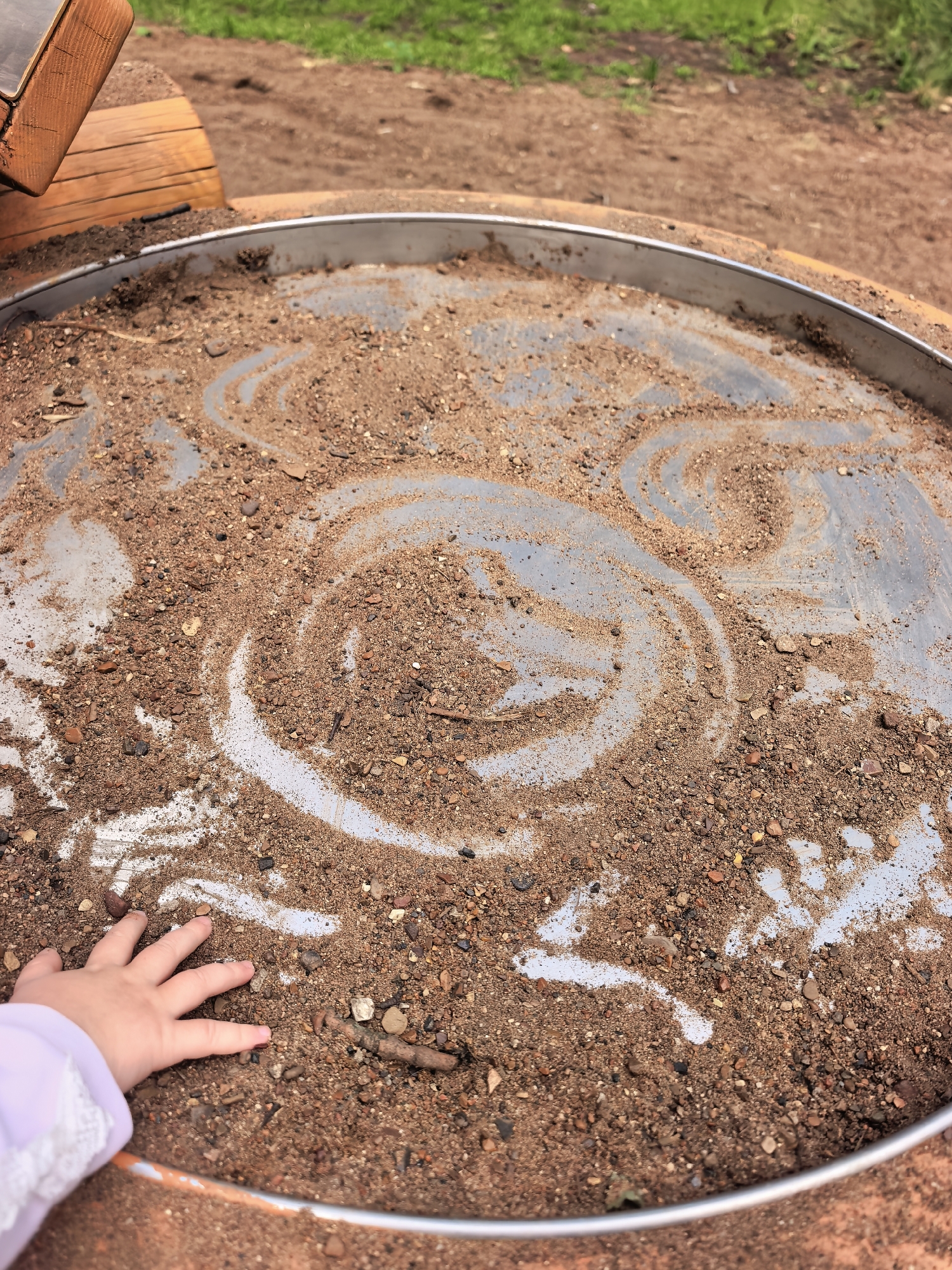 A child's hand with a light purple sleeve reaching into a shallow, circular metal bin filled with sand and small pebbles. The sand has been disturbed, creating swirling patterns. The bin is situated outdoors on a dirt and grass area. The image is taken from a slightly elevated, downward angle, focusing on the sand and the child's hand. There are no specific identifiable landmarks indicating the city of Sterlitamak, Russia.
