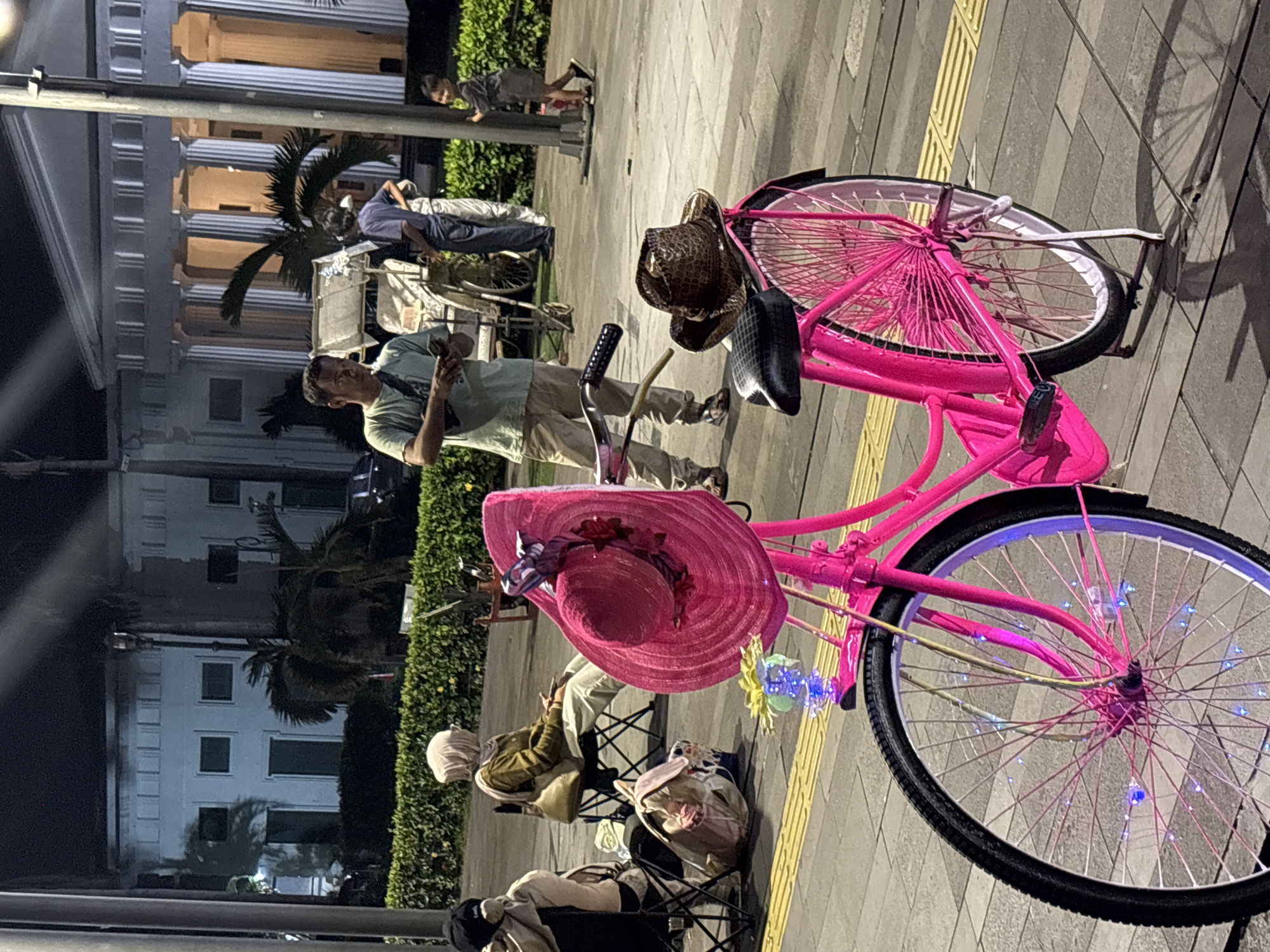 The image depicts a bright pink bicycle with a large pink straw hat adorned with a ribbon and artificial flowers on its handlebars, parked on a sidewalk at night. The bicycle also has small blue lights wrapped around its spokes. A brown, woven cowboy hat rests on the rear rack of the bicycle. In the background, a man in a light-colored t-shirt and tan pants is looking at his phone, standing near a pedicab. Other people are visible sitting on chairs in the mid-ground, including a woman wearing a beige hijab and a tan jacket, sitting in a foldable chair. Further in the background, a colonial-style building with a colonnade and a few palm trees are illuminated. The scene appears to be in front of a historical building, possibly the Fatahillah Square in Kota Tua, West Jakarta, Indonesia, given the architectural style and the presence of a pedicab, which are common in the area. The ground is paved with interlocking bricks and textured tactile paving.