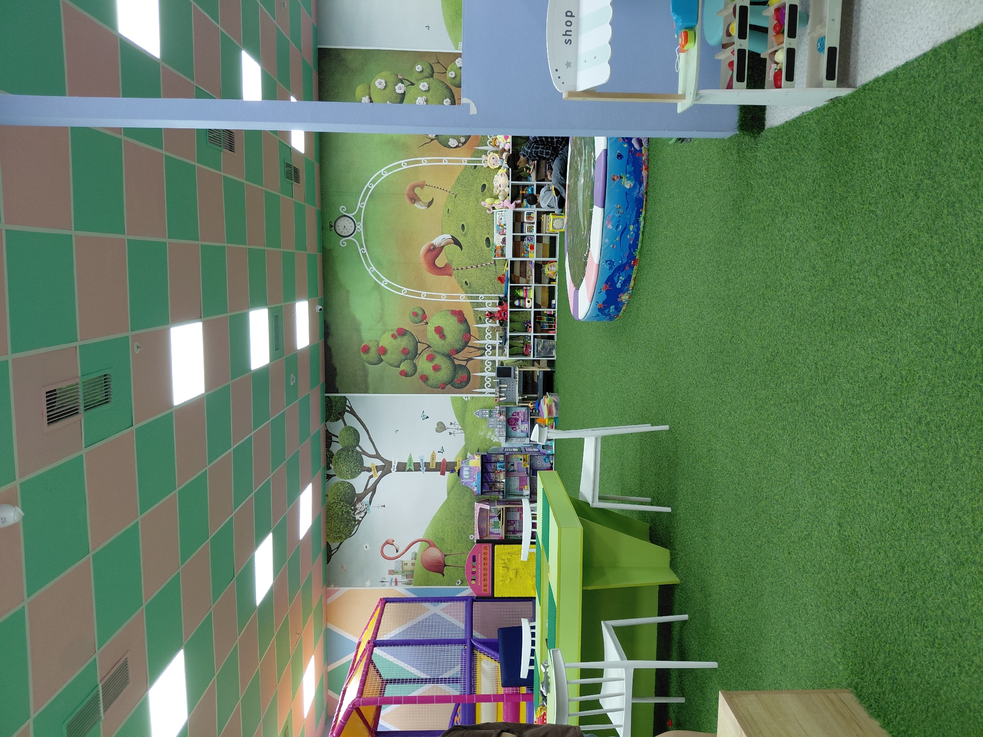The image depicts the interior of a children's play area, likely a playroom or indoor playground. The floor is covered with artificial green turf. In the foreground, a bright green, multi-sided table with a Lego-like surface is set up, surrounded by white chairs. To the left, part of a wooden cubby or structure is visible.
Further back, a large, colorful soft play structure with purple, yellow, and blue sections is present. Adjacent to this structure are shelves filled with toys and games. A large mural adorns the back wall, featuring whimsical drawings of trees, flamingos, and a whimsical landscape with a clock at the top.
In the middle ground, a circular, inflatable pool with a colorful border depicting marine life is visible. A man and a child are seated inside the pool. To the right, a small, white play shop counter with shelves displaying toy food items is positioned. The ceiling is a checkerboard pattern of light pink and green tiles, with fluorescent lighting panels. The scene is brightly lit and appears to be a vibrant and engaging environment for children. The location is identified as Karaganda, Kazakhstan, suggesting this is a specific establishment within the city.