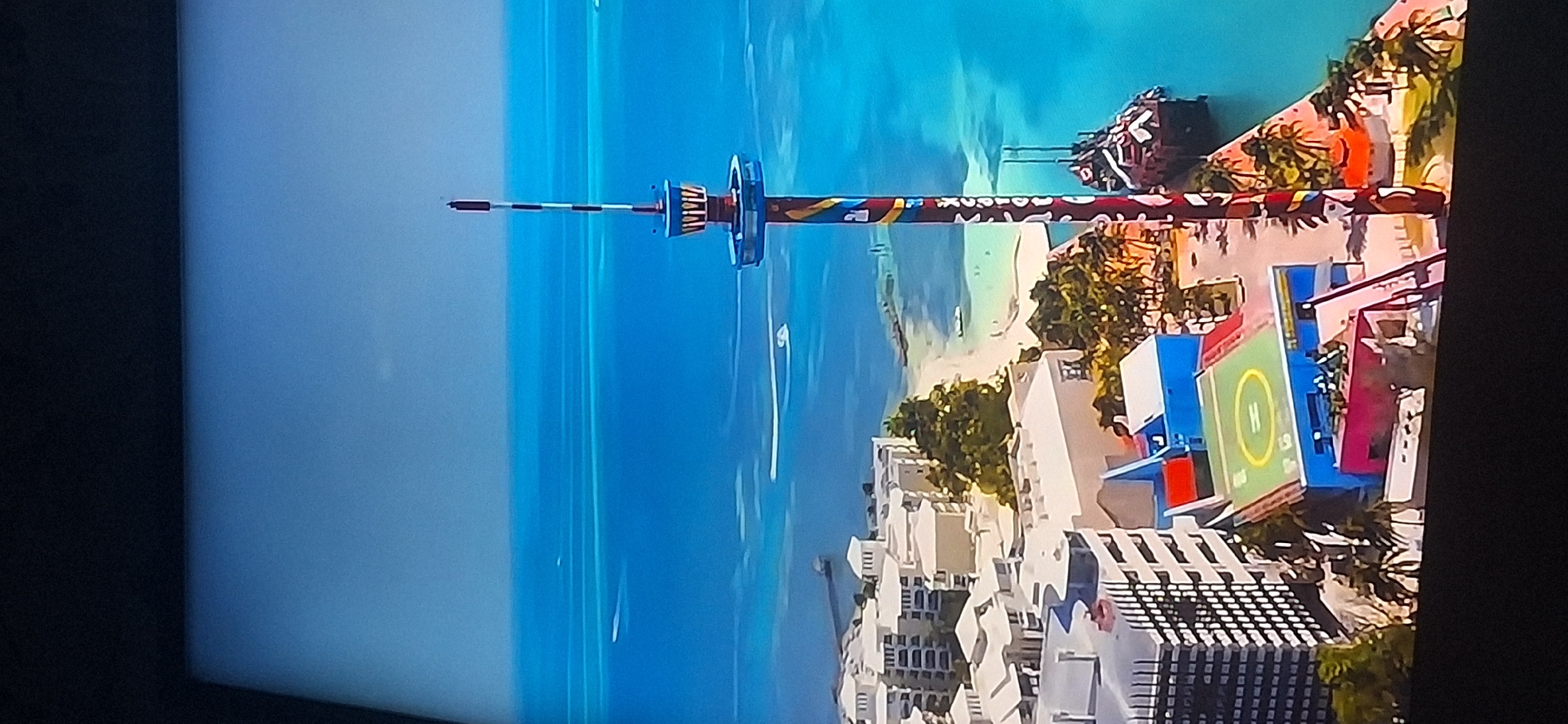 The image is an aerial view of a coastal resort area with a prominent observation tower. The tower is tall and slender, with a colorful, patterned shaft and a circular observation deck near the top. It is situated next to a body of calm, turquoise water, likely the ocean or a large bay.
In the foreground, there are several multi-story buildings, appearing to be hotels or apartment complexes, with white facades and balconies. Some of these buildings are clustered together, forming a dense urban or resort development. A helipad is visible on top of one of the buildings, marked with a yellow "H" inside a circle. There are also smaller, colorful buildings with red and blue roofs. Palm trees and other tropical vegetation are scattered throughout the area, adding to the resort atmosphere.
The water is a vibrant blue, with lighter shades indicating shallower areas or sandbars. There are a few boats visible in the water, leaving white wakes behind them. A pier or jetty extends from the shore into the water in the distance. The sky is a pale blue and appears clear.
The image does not depict any people. The scene is a sunny, tropical environment, possibly a tourist destination.
There is no indication that this location is in Bila Tserkva, Ukraine. Bila Tserkva is an inland city and does not have a coastline or such a resort-like setting.