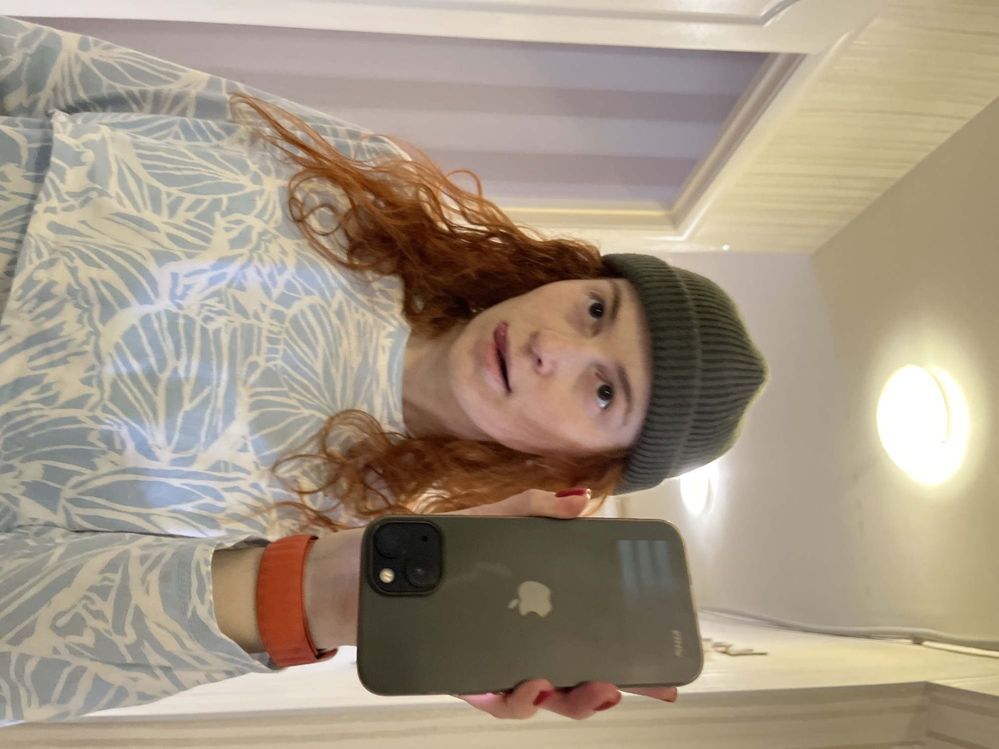 An indoor mirror selfie taken in Chaikovsky, Russia, features a person looking directly at the camera. The subject has long, wavy reddish-brown hair and wears a light blue long-sleeved shirt with a white leaf-like pattern. A dark green knit beanie covers the top of their head. Their expression is neutral, with lips slightly parted, and their eyes are wide.
In their right hand, the person holds a dark gray iPhone, with its back facing the viewer. The prominent Apple logo is visible on the phone's case, which also bears the small text "fitte" near the bottom. An orange band, resembling a smartwatch or fitness tracker, is on their left wrist. Red nail polish is visible on their fingers.
The background consists of light-colored walls and a ceiling with architectural details, possibly molding or a skylight structure. Two circular, warm-toned wall-mounted lights are visible to the right, providing illumination. A white wire runs along the base of the wall. The scene suggests a modern, well-lit interior space.