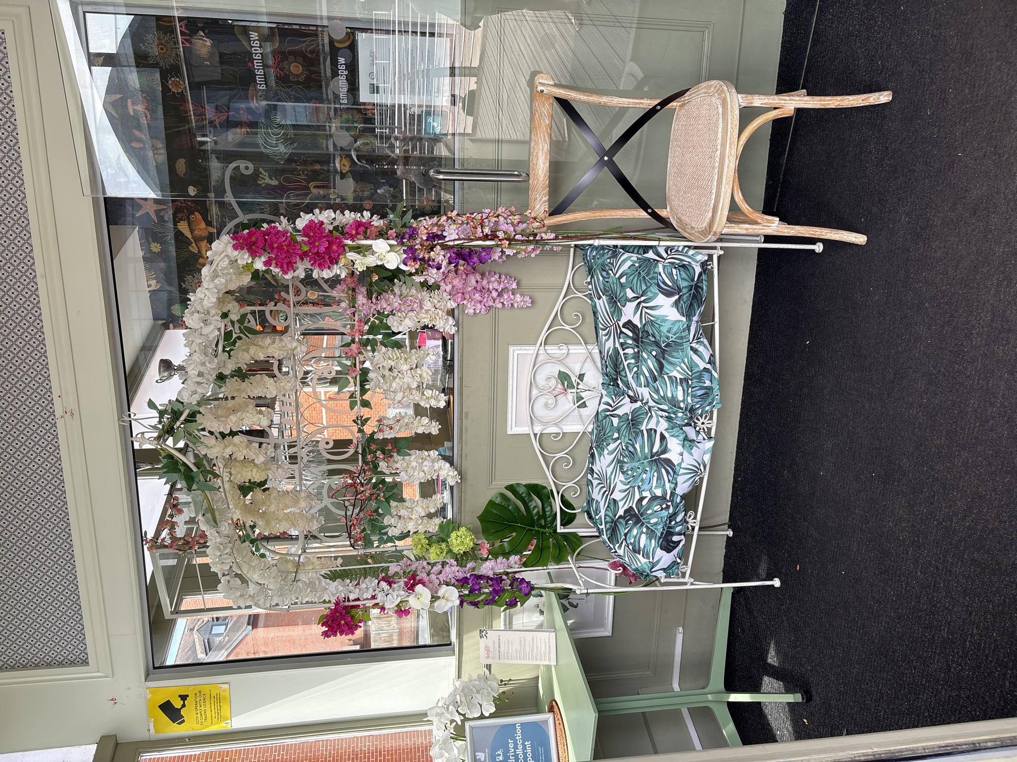 An outdoor scene features a display of artificial flowers and a bench in front of a glass-paneled door. The floral arrangement is a mix of white, pink, and purple blossoms cascading down a white metal structure. A white bench with a tropical leaf-patterned cushion is placed next to the flowers. A light-colored wooden chair with a black criss-cross back is positioned behind the display. The setting appears to be a storefront, with the interior visible through the glass. Inside, there are shelves and decor, including what looks like artwork and possibly merchandise.
A yellow and black sign is affixed to the glass, which reads " CCTV IN OPERATION " and " WE OPERATE WITH OUR TRADING LICENCE ". Another sign on a green stand inside the store is partially visible, with some text. A blue sign on a green table inside the store reads " DELIVERY DRIVER COLLECTION POINT ". The ground in front of the display is covered in a dark carpet. The time of day appears to be daytime, with sunlight casting shadows. The overall impression is a welcoming and decorative display outside a business.