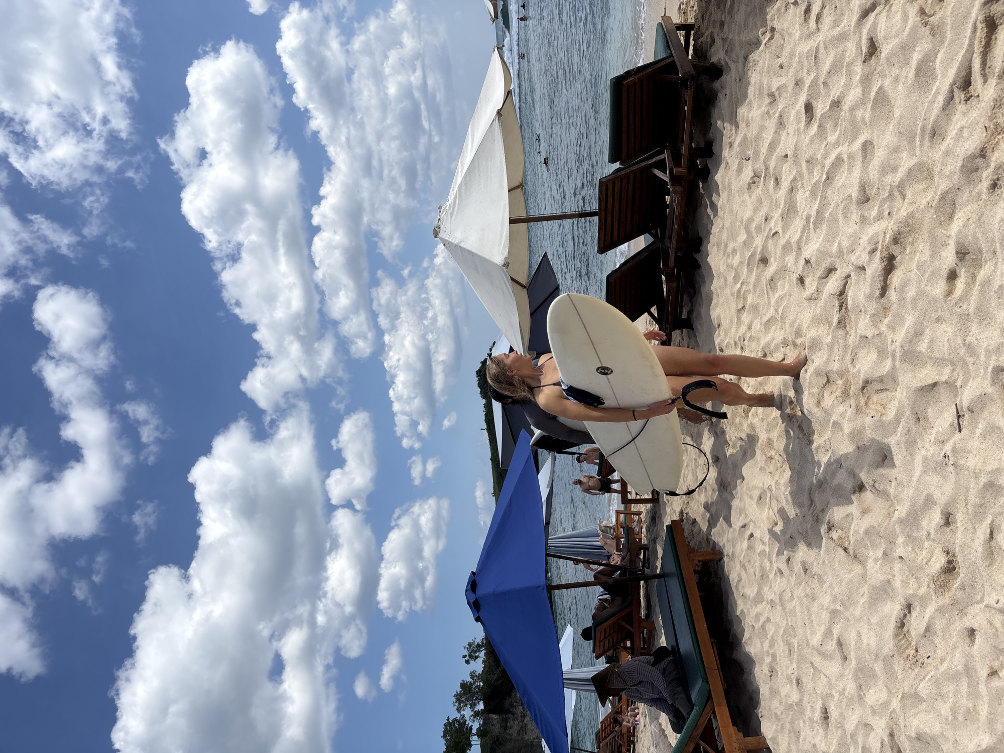 The image depicts a sunlit beach scene in Kuta Selatan, Indonesia, under a bright blue sky with scattered white cumulus clouds. In the foreground, a fair-skinned woman with light hair, wearing a dark bikini top and bottoms, walks across light-colored sand from left to right, carrying a white shortboard surfboard under her left arm. She looks towards the ocean. Distinct shadows of the woman and other beach structures are cast on the sand.
To her left, a large dark blue beach umbrella shades multiple wooden lounge chairs with green cushions. One individual on a lounge chair wears a wide-brimmed straw hat and a striped shirt. To her right, a large white beach umbrella also shades several unoccupied wooden lounge chairs. The midground shows more lounge chairs, some occupied, extending towards the water's edge. Several people are visible in the shallow ocean and further out, with approximately 5-7 individuals, some on surfboards, suggesting surfing activity. Small waves are breaking in the distance. On the far left, a rocky cliff or elevated landmass with sparse vegetation is visible.