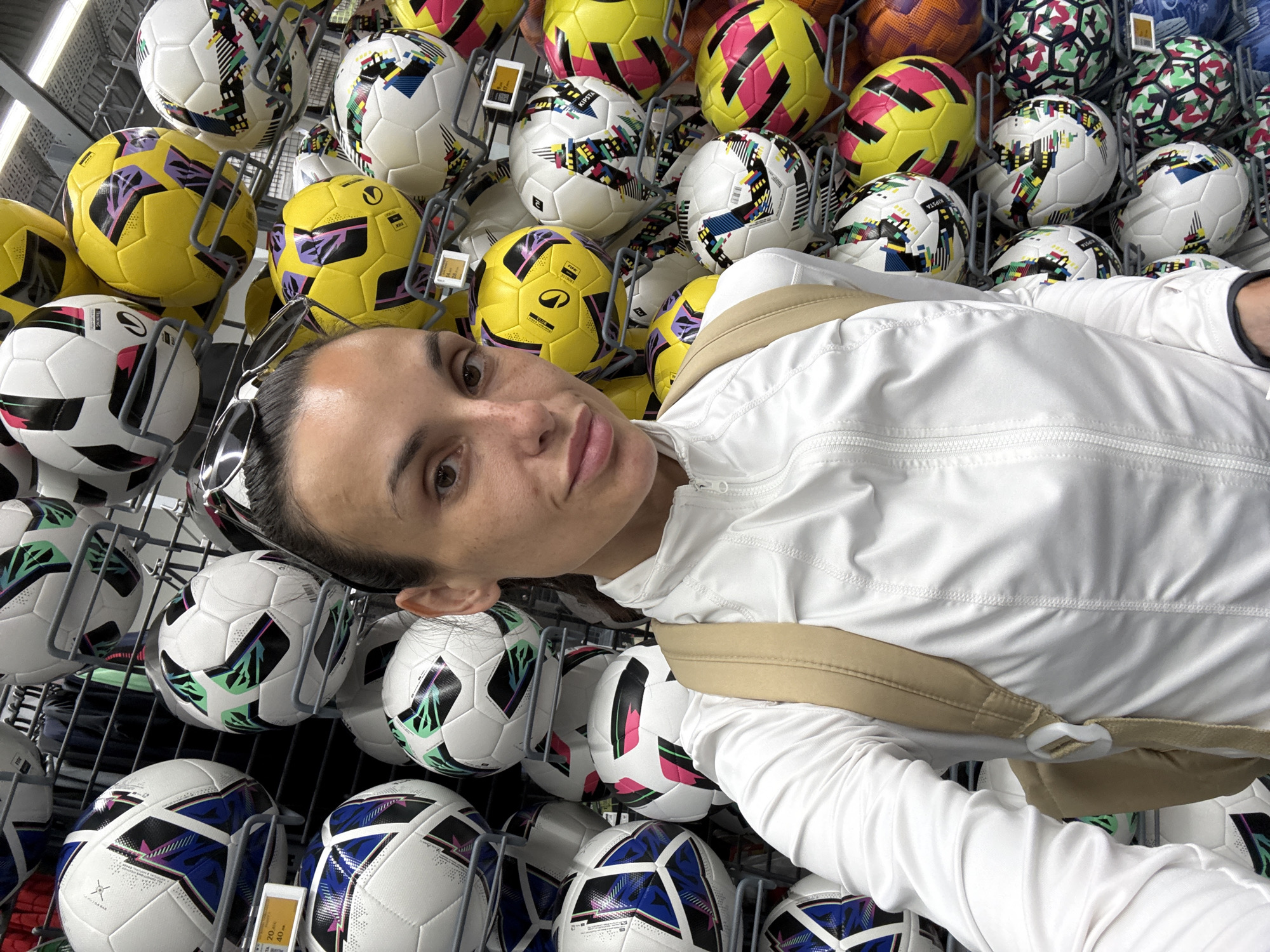 A woman with dark hair and sunglasses on her head is taking a selfie in front of a display of soccer balls. She is wearing a white jacket and a tan backpack. The soccer balls are of various colors, including white, yellow, green, and pink. The display is set up on metal shelves. The image appears to be taken indoors, possibly in a sporting goods store. The location is stated to be the city of Pleven, Bulgaria.