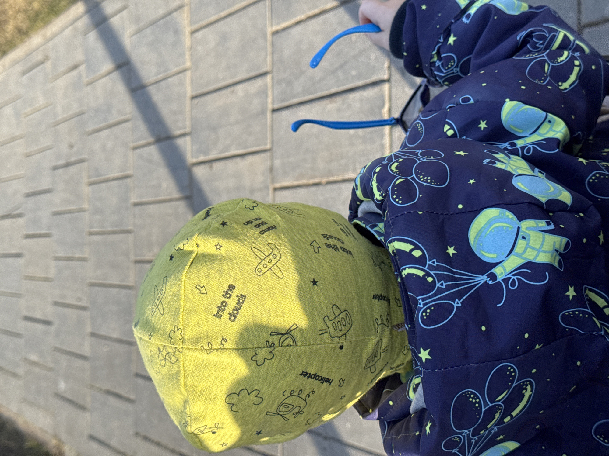 An overhead outdoor shot depicts a young child wearing a dark blue spacesuit-patterned jacket with lime green details, holding blue eyeglass frames. The child's head is covered by a chartreuse cap adorned with airplane and cloud graphics. They are standing on a grey paved area composed of rectangular blocks. The location is not stated, nor is there any information to indicate that the image was taken in the city of Izhevsk, Russia.