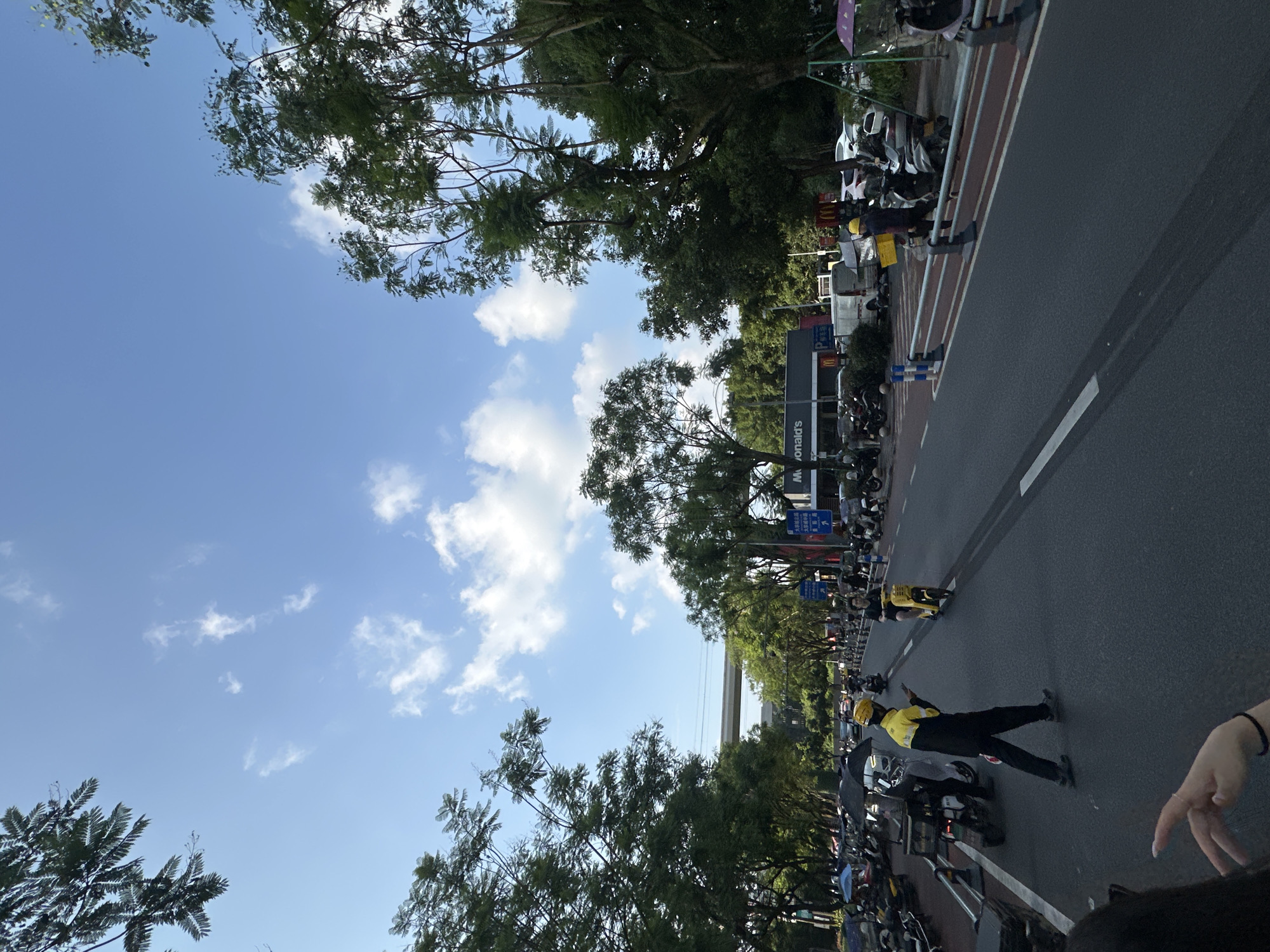 The image depicts a street scene in the Chongqing High-tech Zone, China. The sky is a clear blue with scattered white clouds. On the left side of the road, numerous parked motorcycles and scooters are visible, some with umbrellas covering them. A person in a yellow and black uniform, likely a delivery worker, is walking across the street, looking at their phone. To the right of the road, more motorcycles and parked cars are present, along with a McDonald's restaurant. Trees line both sides of the street, and an elevated road or bridge structure is visible in the background. The scene appears to be in a moderately busy urban environment with ongoing traffic and pedestrian activity.