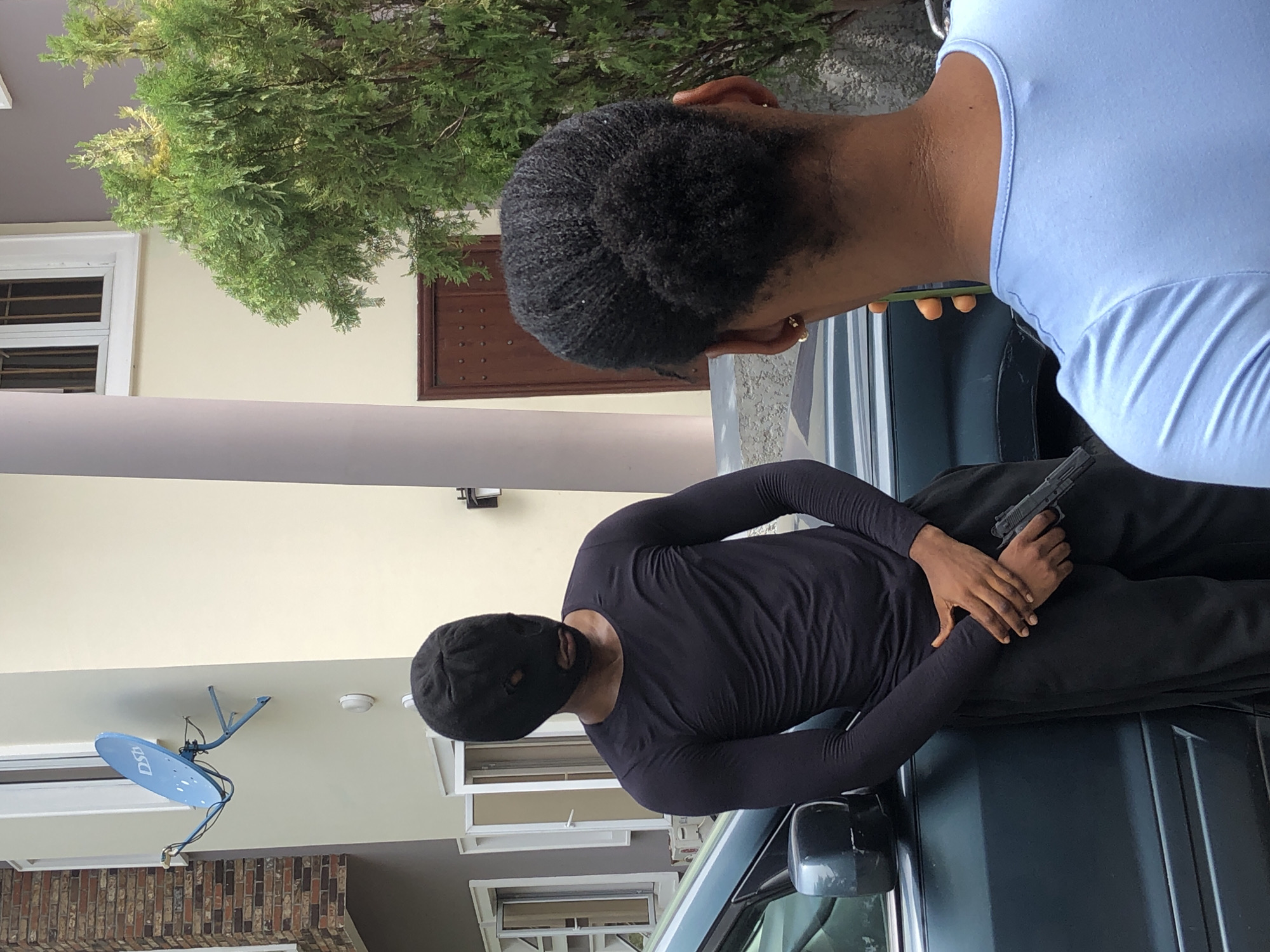 This image captures an outdoor scene, appearing to be in front of a residential building, with two individuals and a vehicle. The photograph is taken from a slightly elevated perspective.
In the foreground, on the right side, is the back of a person with short, dark, curly hair, wearing a light blue top. Their head is tilted downwards, suggesting they are looking at or interacting with the person on the car.
The central subject is a person dressed entirely in black, including a long-sleeved top and trousers. Their face is covered by a black balaclava or ski mask, obscuring their identity, with visible eyeholes. This individual is lying horizontally on the hood or roof of a dark-colored vehicle, which appears to be an SUV or car, with a visible side mirror. They are holding a dark-colored handgun with both hands, resting it on their midsection. Their posture appears posed and static.
The background features a light-colored building with cream or light yellow walls. Visible architectural elements include white-framed windows, light grey or beige pillars, and a section of a brown brick wall. A large, green tree or bush with dense foliage extends across the upper left portion of the frame. A dark brown door with decorative circular details is visible further back. A light blue satellite dish, bearing the text "DSTV," is attached to the building on the left side.
The scene appears to be set during daytime under bright conditions. The presence of the "DSTV" satellite dish suggests a location in Africa. There are no overt signs of active movement or strong emotions from the visible subjects; the scene has a staged quality.