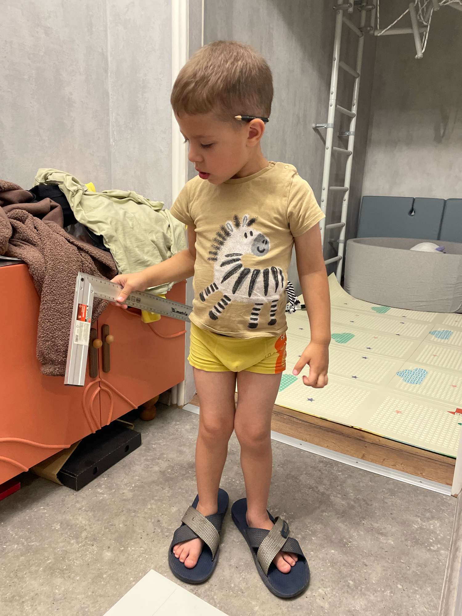 A young boy, estimated to be around 4-5 years old, stands in a room. He has short brown hair and is wearing a light brown t-shirt with a zebra print and yellow underwear. He is holding a silver L-shaped ruler, examining it. The room appears to be a child's play area or bedroom. To the left, there is a bright orange cabinet with wooden knobs, piled with clothes. Behind the boy, a grey wall is visible, with a children's climbing frame attached. To the right, there is a padded play mat on the floor, and a grey soft play area. The floor is concrete. There is no discernible evidence to place this image in Talagi, Russia.