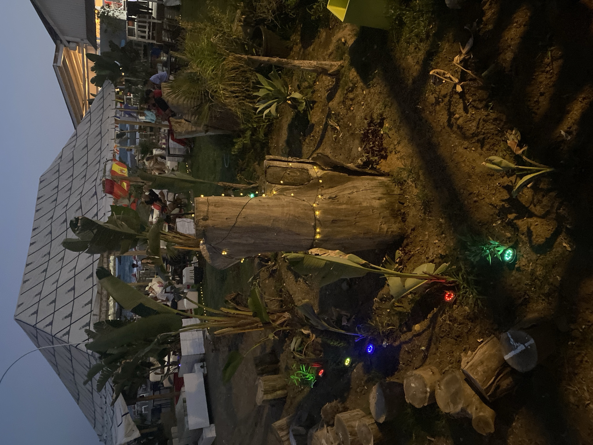 The image depicts an outdoor seating area at dusk, likely a restaurant or bar, situated near the beach in Estepona, Spain. The foreground features a garden with a large tree stump adorned with string lights, surrounded by cut logs and various plants. Several small, colored lights illuminate the ground around the garden. In the background, a large, open-sided structure with a distinctive geometric roof provides shade for patrons seated at tables. Beyond this structure, a glimpse of the beach and the sea is visible. The overall atmosphere is relaxed and inviting, with people enjoying the evening outdoors.