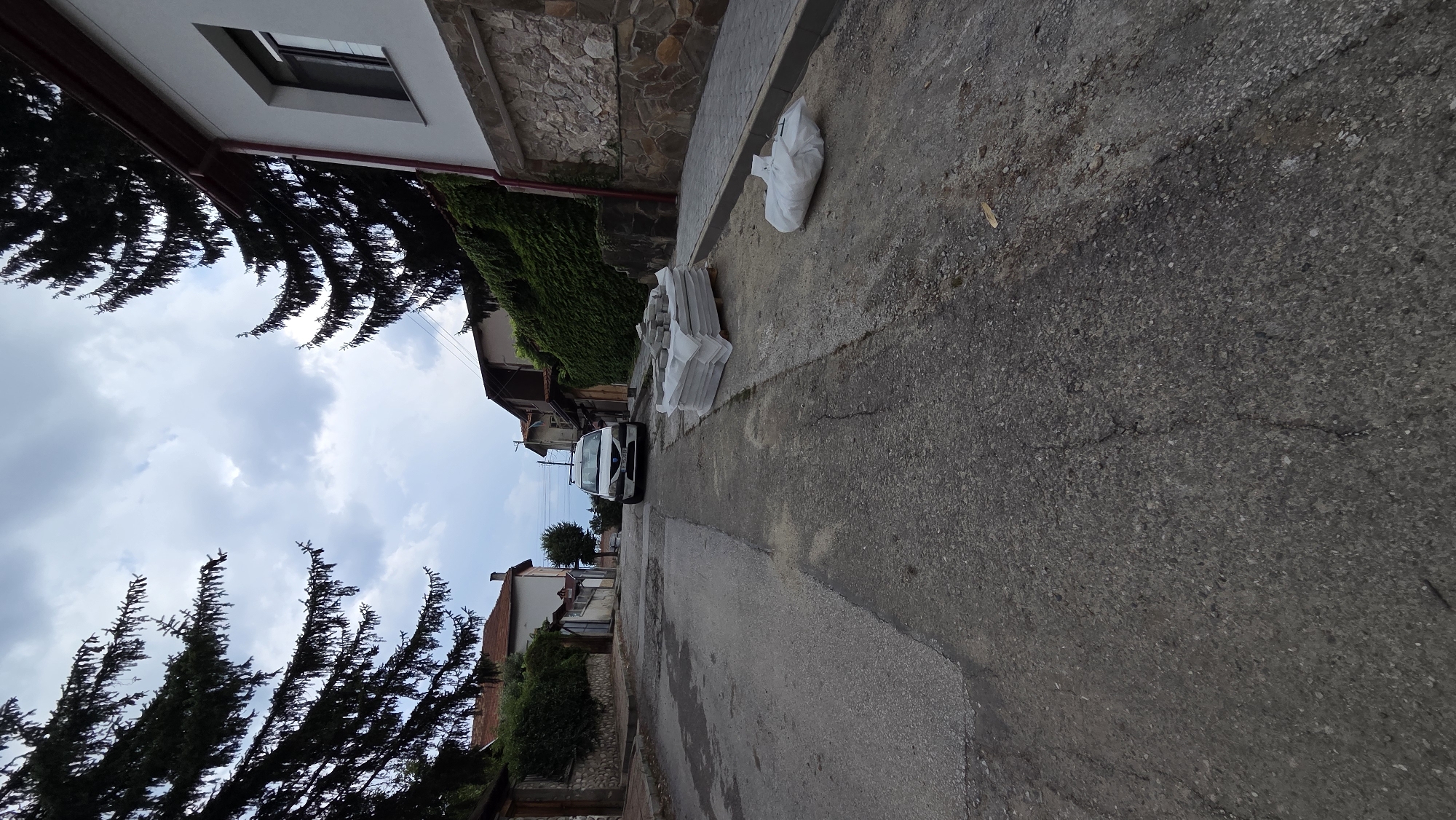 The image captures a street scene in the city of Bania, Bulgaria. The perspective is low, looking up a paved street that appears to be undergoing some form of construction or repair. In the middle ground, a white van is parked facing uphill. To the right of the van, on the street, there is a pallet stacked with grey paving stones, covered in white plastic sheeting. Further to the right, on the side of the street, a large white bag is also present. The street is flanked by houses with traditional architecture, some with visible stone foundations and tiled roofs. Evergreen trees with dark foliage are prominent on either side of the street, framing the view towards the sky, which is overcast with grey clouds. The ground of the street is asphalt with visible cracks and patches.