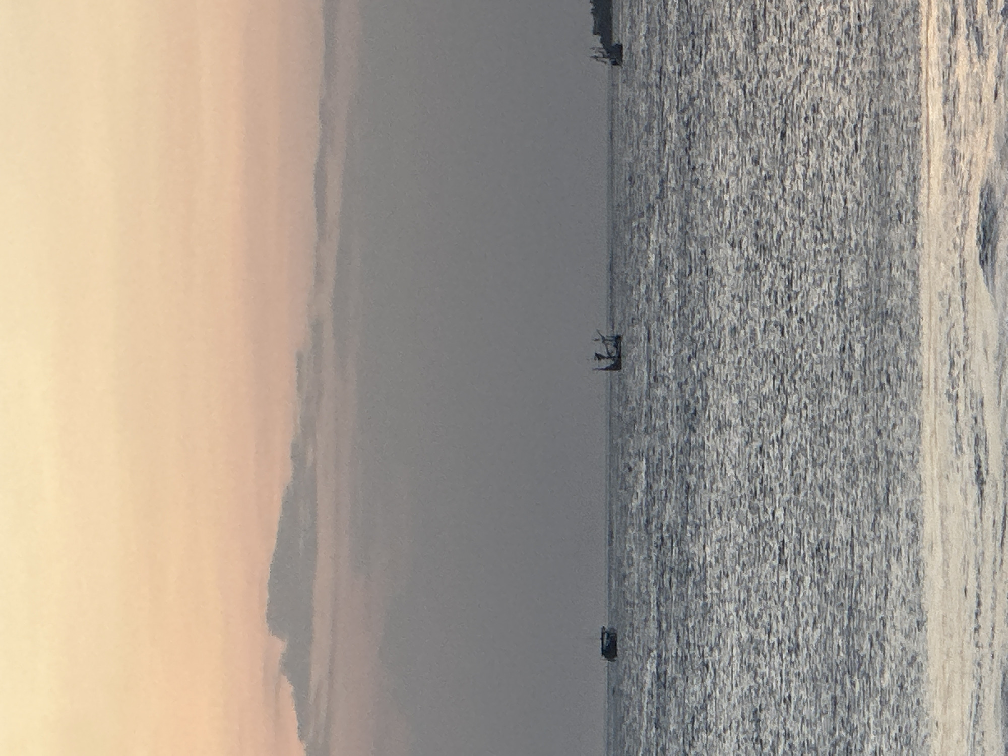 This is a photograph of the ocean with a soft, hazy sky. The image is oriented vertically, with the sky on the left and the water on the right. The sky is a gradient of pale yellow and peach, suggesting it might be sunrise or sunset. The water is a deep gray-blue and appears choppy, with whitecaps visible. There are several small objects in the water, which appear to be boats. One boat in the middle distance has what looks like two people in it. Another boat is closer to the foreground. The horizon line is faint and blends with the hazy sky. The overall mood is tranquil and serene, characteristic of a quiet moment by the sea. Given the location context of Kuta, Indonesia, this scene likely depicts a view of the Indian Ocean. The time of day is suggested to be either early morning or late evening due to the soft lighting. The weather appears to be clear, though hazy.