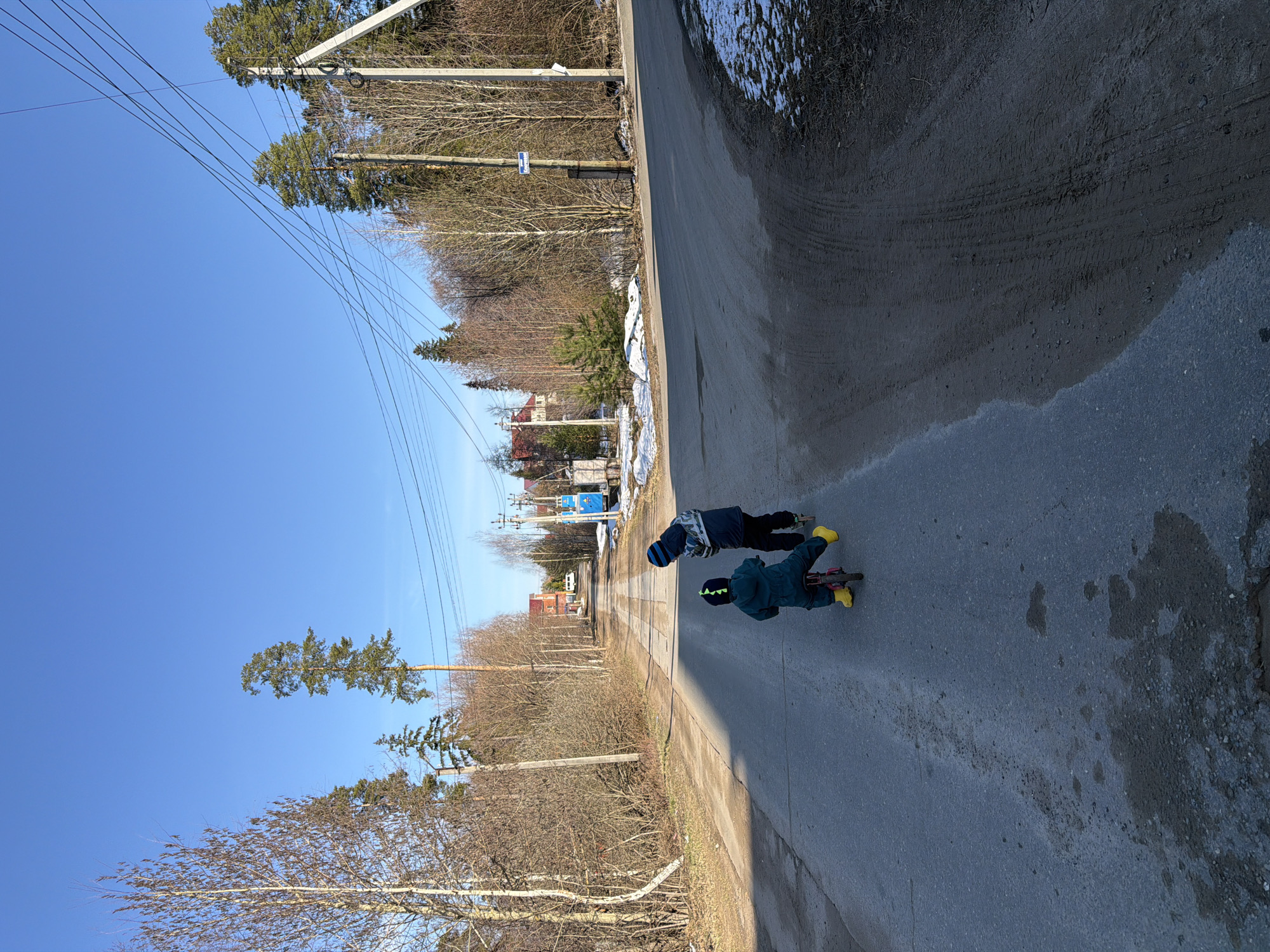 Two children are on a street in Chaikovsky, Russia, riding bicycles. The street is asphalt and shows evidence of recent water runoff. Trees line both sides of the road, some still retaining snow. Overhead, power lines stretch across the sky. Buildings and infrastructure are visible in the distance.