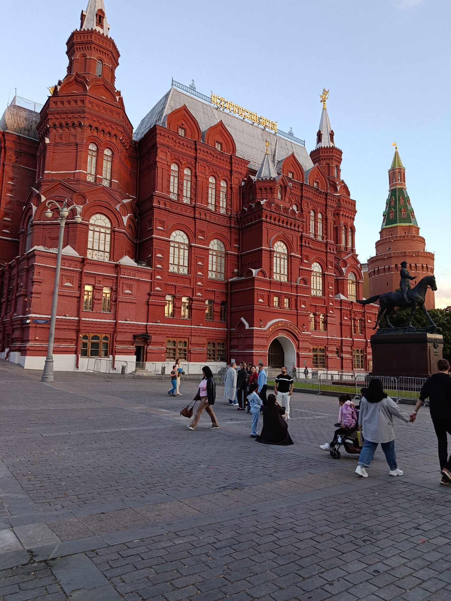 The image depicts a lively public square in front of a grand, historical red brick building under a clear, late afternoon or early evening sky. The central subject is a multi-turreted structure with intricate white architectural detailing around its numerous arched windows. Gold Cyrillic text on the roof clearly spells out "ГОСУДАРСТВЕННЫЙ ИСТОРИЧЕСКИЙ МУЗЕЙ" (State Historical Museum), serving as a significant location cue.
In the midground, an equestrian statue of a man on horseback is prominently positioned to the right of the main building. Further in the background on the right, another red brick tower with a distinct green conical roof is visible. The foreground and midground consist of a paved plaza where numerous people are casually walking and congregating. They are dressed in modern, diverse attire, with individuals and small groups moving across the square. One person is seen pushing a child in a stroller-like device. The atmosphere appears relaxed and calm, reflecting everyday urban activity.
Notable details include the warm, low-angle light suggesting the time of day is late afternoon or early evening. The sky is a pale blue with no clouds, indicating clear weather. A tall, ornate streetlamp stands on the left side of the plaza. Metal barriers are placed along the base of the main building, separating it from the pedestrians. Another partially visible sign on the building's facade, also in Cyrillic, can be seen near the streetlamp. The scene captures a blend of impressive historical architecture and contemporary public life.