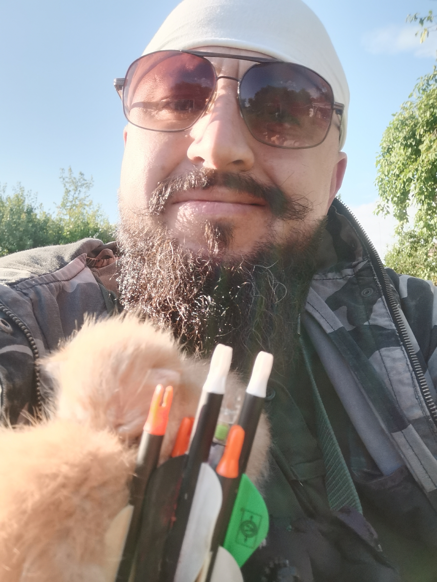 The image shows a close-up, selfie-style shot of a man wearing sunglasses and a white head covering. He has a beard and a mustache. He is holding a small, fluffy, light-colored animal, possibly a kitten, in his left arm. In front of the animal and the man's chest, a group of arrows with orange and white fletching are visible. The man is wearing a camouflage jacket over a lighter-colored shirt.
The setting appears to be outdoors, with a clear blue sky and greenery in the background, suggesting it is daytime. The lighting indicates it could be a sunny day. The man's expression is neutral, and the overall mood is casual. There is no visible text in the image. The provided location context places this scene in Semypolky, Ukraine.