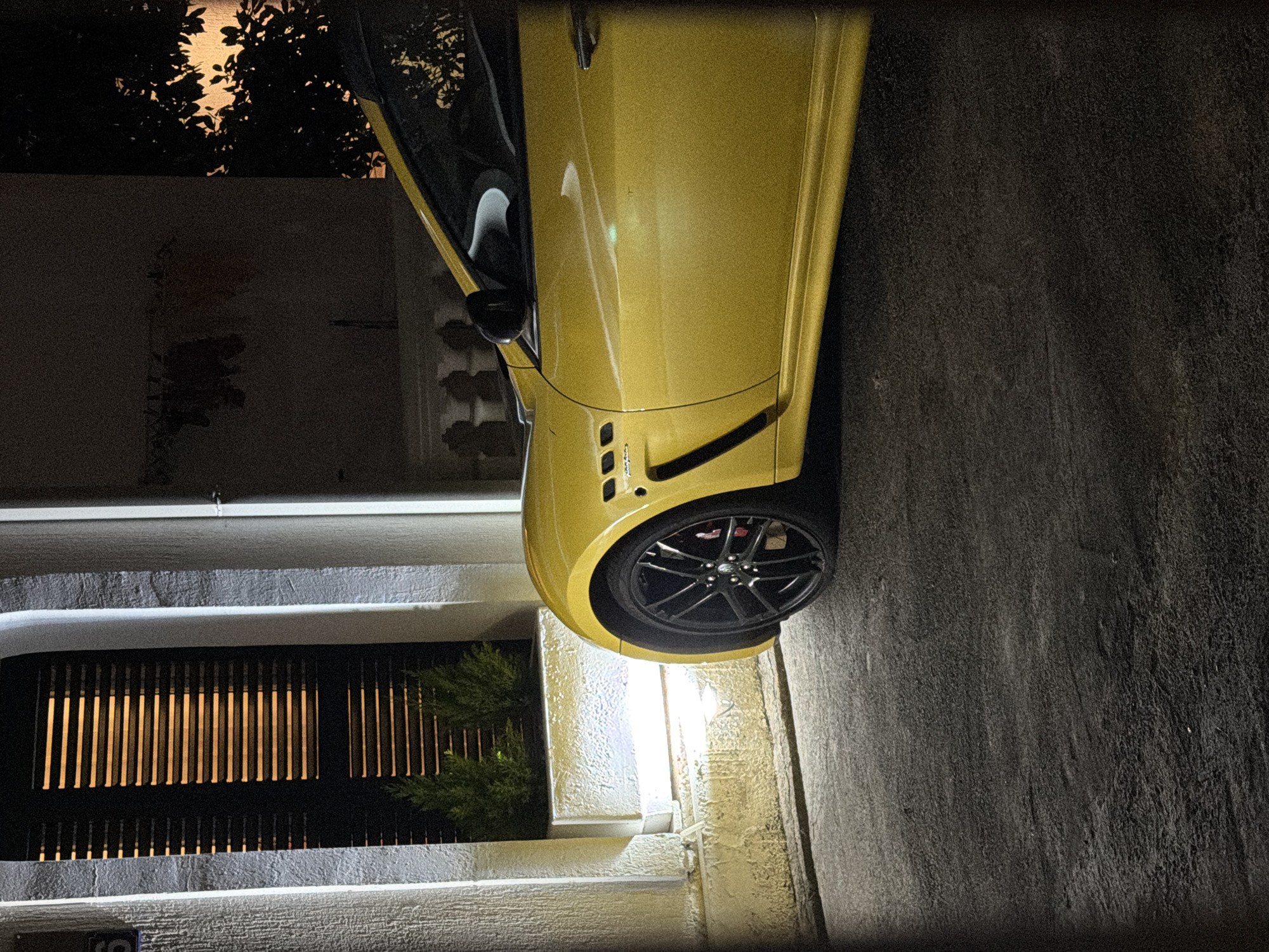 The image shows a close-up of the front passenger side of a yellow sports car parked on an asphalt surface at night. The car's wheel, fender, and a portion of the door are visible. The car is positioned in front of a building with a textured, light-colored facade. To the left of the car, a window with horizontal blinds is illuminated from within, casting striped patterns. Two small, green evergreen plants are visible in a planter box beneath the window. White vertical lights are positioned on either side of the window area. The background is dark, with foliage visible on the right side, backlit by an unseen light source. The overall scene is dimly lit, typical of an outdoor urban setting at night. The specific location in Budva, Montenegro, is not explicitly indicated by any visible landmarks or text.