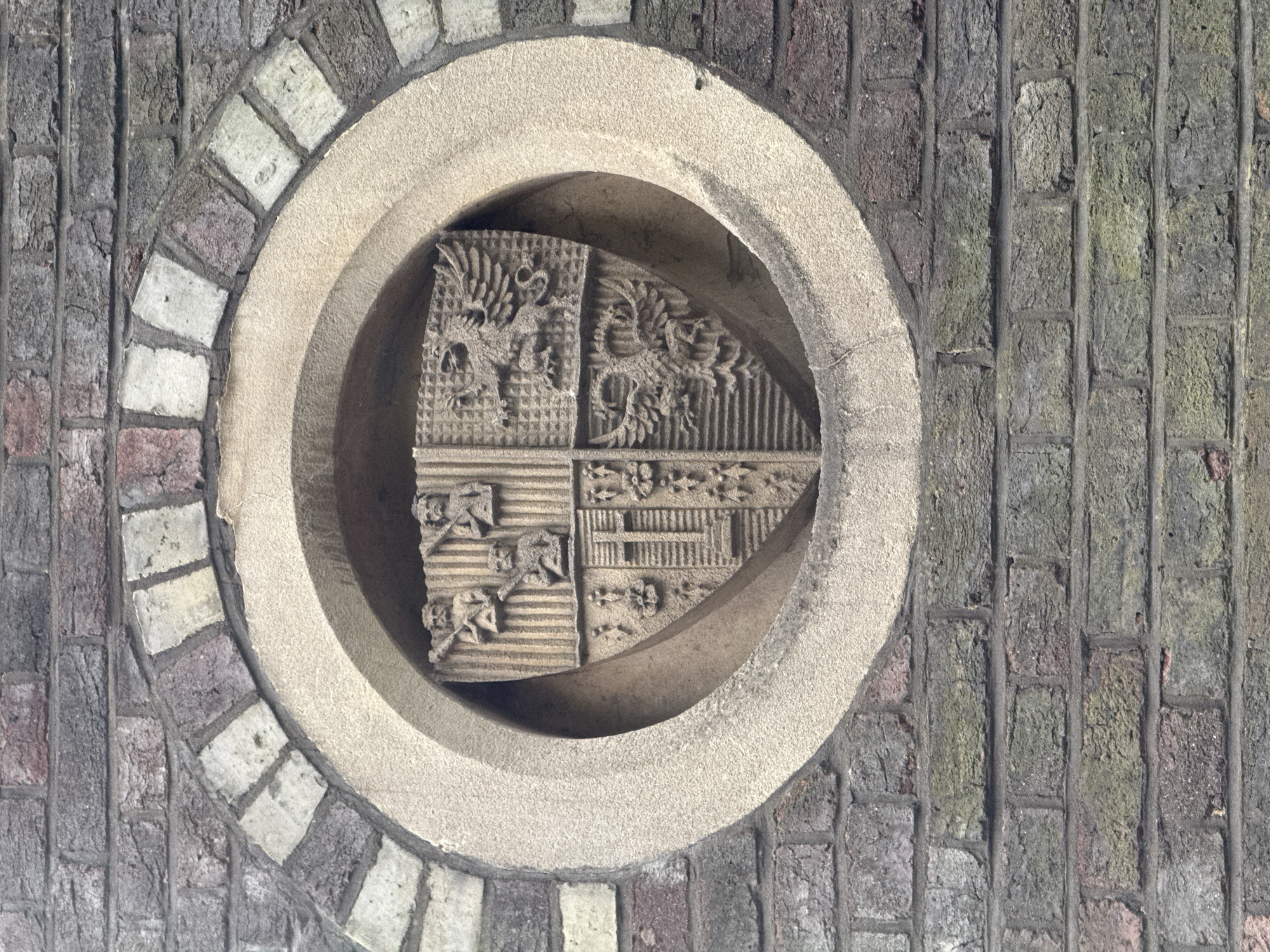 A stone carving, seemingly a coat of arms, is set within a circular niche on a brick wall. The wall has a multi-toned brick arrangement, including gray, purple, red, and light stone-colored bricks. The circular niche surrounding the carving is formed from a lighter stone than the brick wall. The carving itself is a complex design, divided into four distinct quarters. Two of the quarters depict what appear to be dragon-like figures, while the other two display abstract and geometric patterns. The texture of the stone is rough and weathered. The image appears to be taken in London, United Kingdom.
