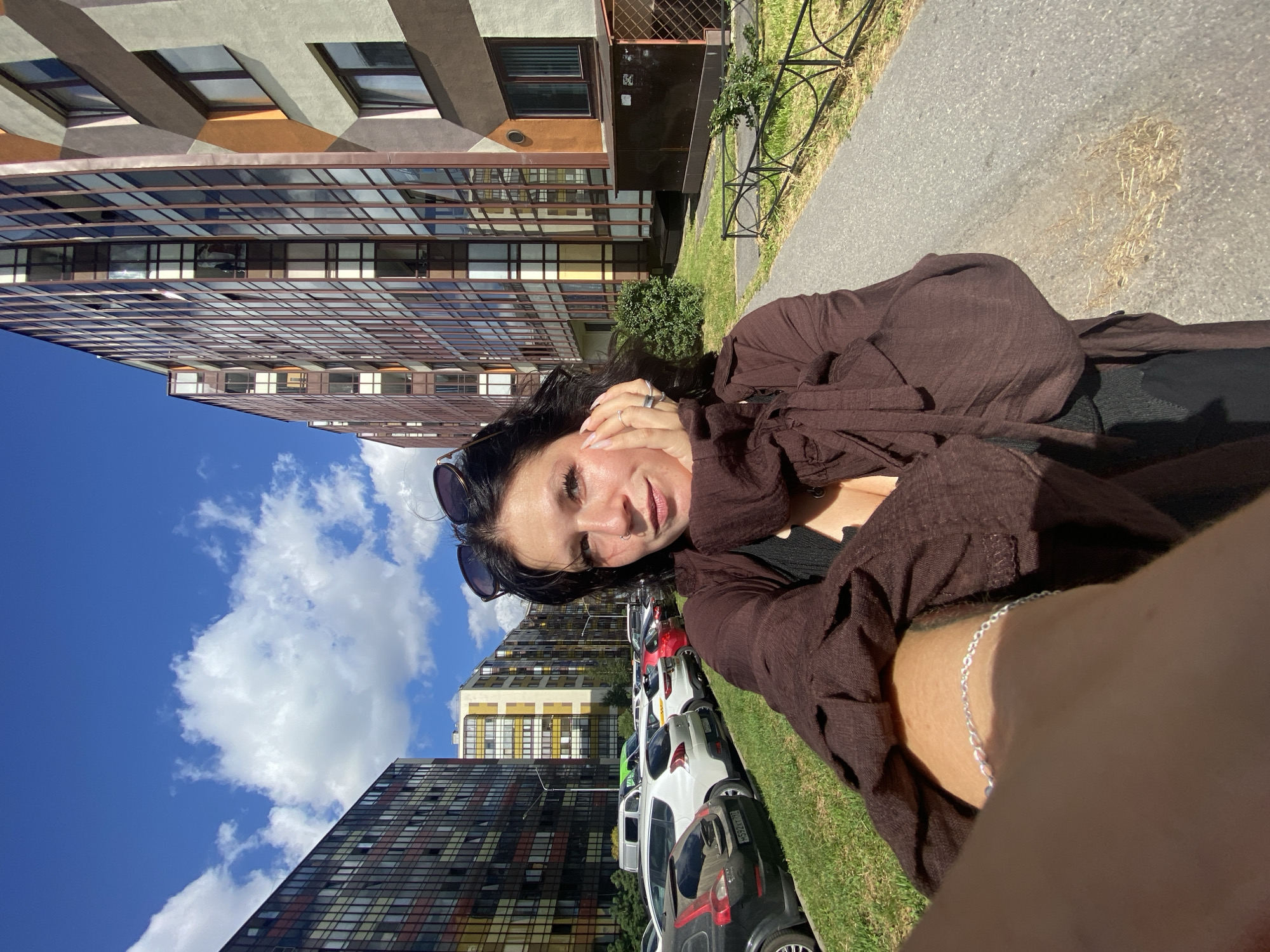 A young woman is taking a selfie outdoors in Kudrovo, Russia. She has long dark hair, is wearing sunglasses on her head, and a brown shirt. Her right hand is resting on her cheek. In the background, there are modern apartment buildings with colorful facades, a parking lot with cars, and a clear blue sky with scattered clouds. The woman is standing on a paved walkway next to a grassy area.