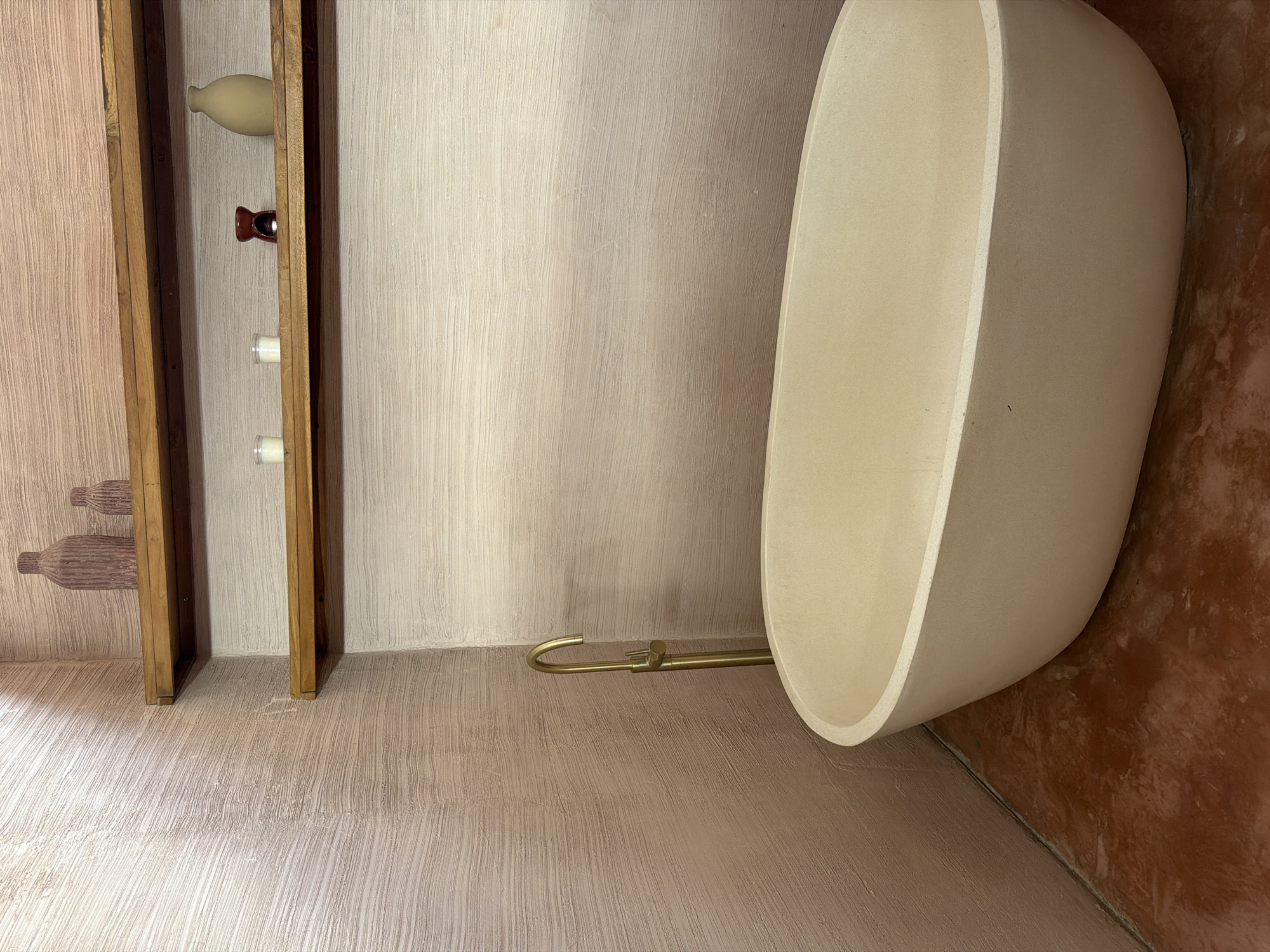 This is an interior shot of a bathroom in North Kuta, Indonesia. The bathroom features a freestanding, oval-shaped tub in a light cream color, positioned against a wall with a textured, light beige finish. A brass-colored faucet extends from the wall towards the tub. To the left, a wooden shelf unit is mounted on the wall, displaying decorative items including a light-colored vase, a small, dark red object, and two white candles. The walls and floor share a similar textured finish, giving the space a uniform aesthetic. The lower right corner of the image shows a portion of a darker, reddish-brown wall.