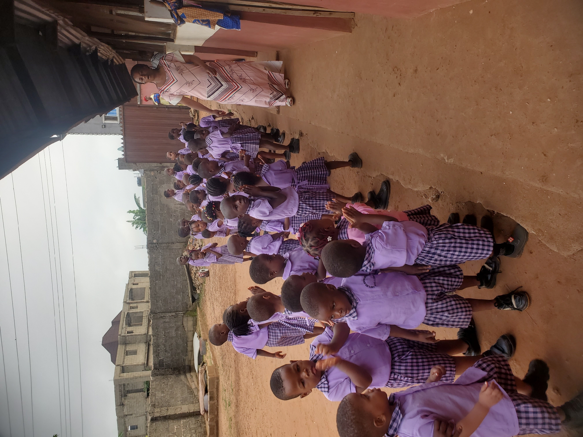 In Ukwu-Abwa, Nigeria, a line of approximately 30 uniformed schoolchildren stands outdoors. They are mostly of African descent and wear light purple shirts and checkered skirts or trousers. Their shoes are black. They appear to be in a prayer formation, with hands held together. A woman in a pink patterned dress stands at the head of the line. The environment appears to be a school yard with buildings and dirt ground.