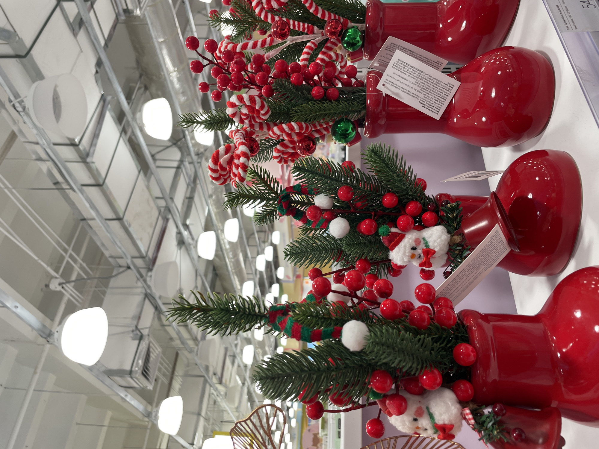 In the city of Davao City, Philippines, inside a retail store, Christmas-themed decorative items are displayed on shelves. The items are primarily artificial pine branches adorned with red berries, white pom-poms, and small snowman figures, all housed in bright red boot-shaped vases. Some arrangements also feature candy canes and green baubles. The background shows the store's industrial ceiling with suspended lighting fixtures and ventilation systems. The overall scene suggests a commercial setting preparing for the holiday season.