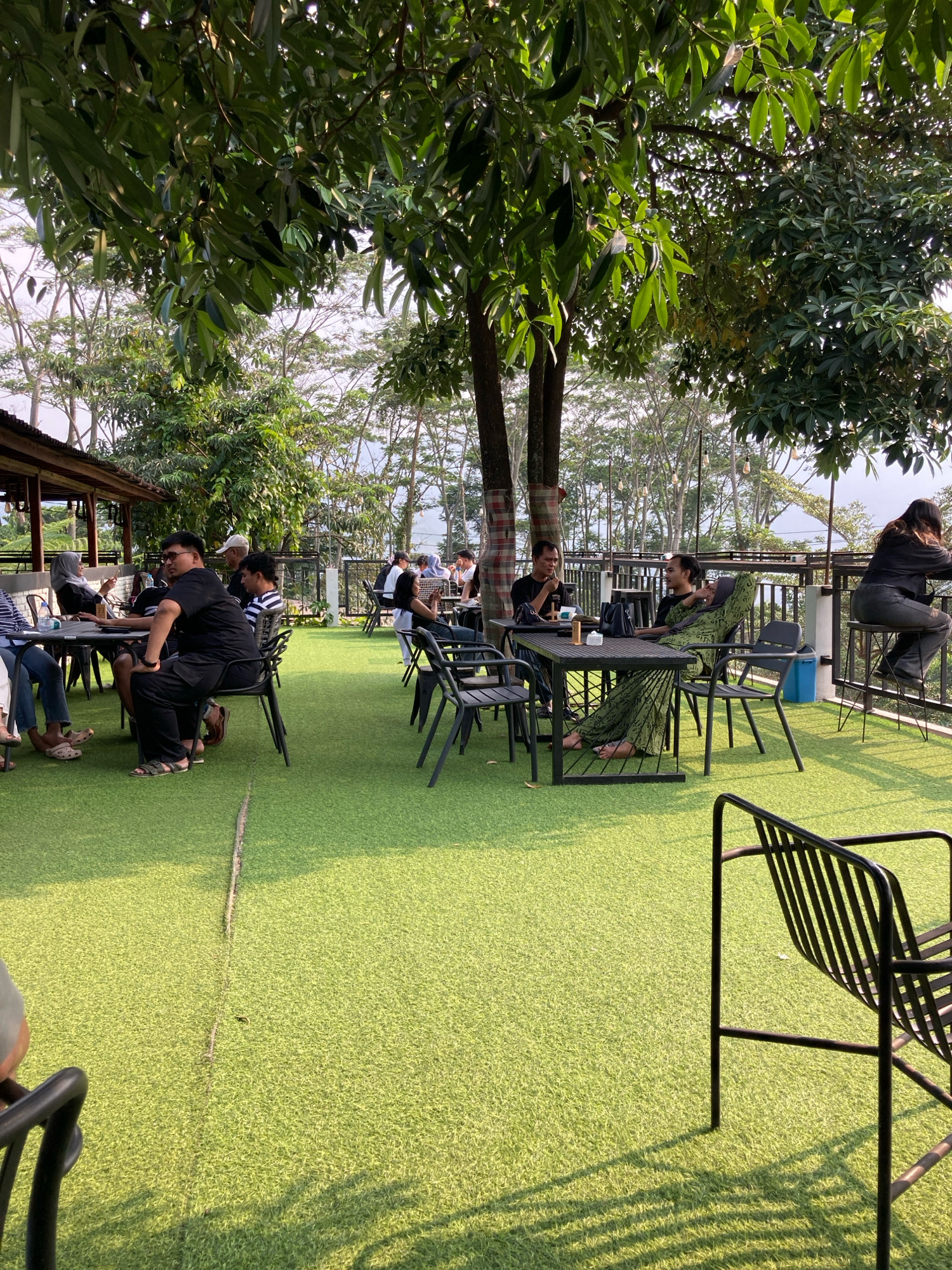 The image captures an outdoor seating area of a cafe in Sukamakmur, Indonesia, characterized by an artificial turf carpet, ample natural light, and lush greenery from overhanging trees. Several groups of people are seated at tables, engaged in various activities. In the foreground, to the left, a person in a black t-shirt and pants is seated at a table, facing left. To their right, a person in a black t-shirt and striped shirt is seated. Further back, a group of individuals are seated at tables, some appear to be in conversation, while others are looking at their phones or drinks. One individual in a green patterned dress is reclined in a chair, looking to the right. To the far right, a person in a black top and grey pants is seated on a stool at a high table, looking away from the camera. The cafe's structure is visible in the background with a rustic roof and railings. The overall ambiance suggests a relaxed setting for dining or socializing.
