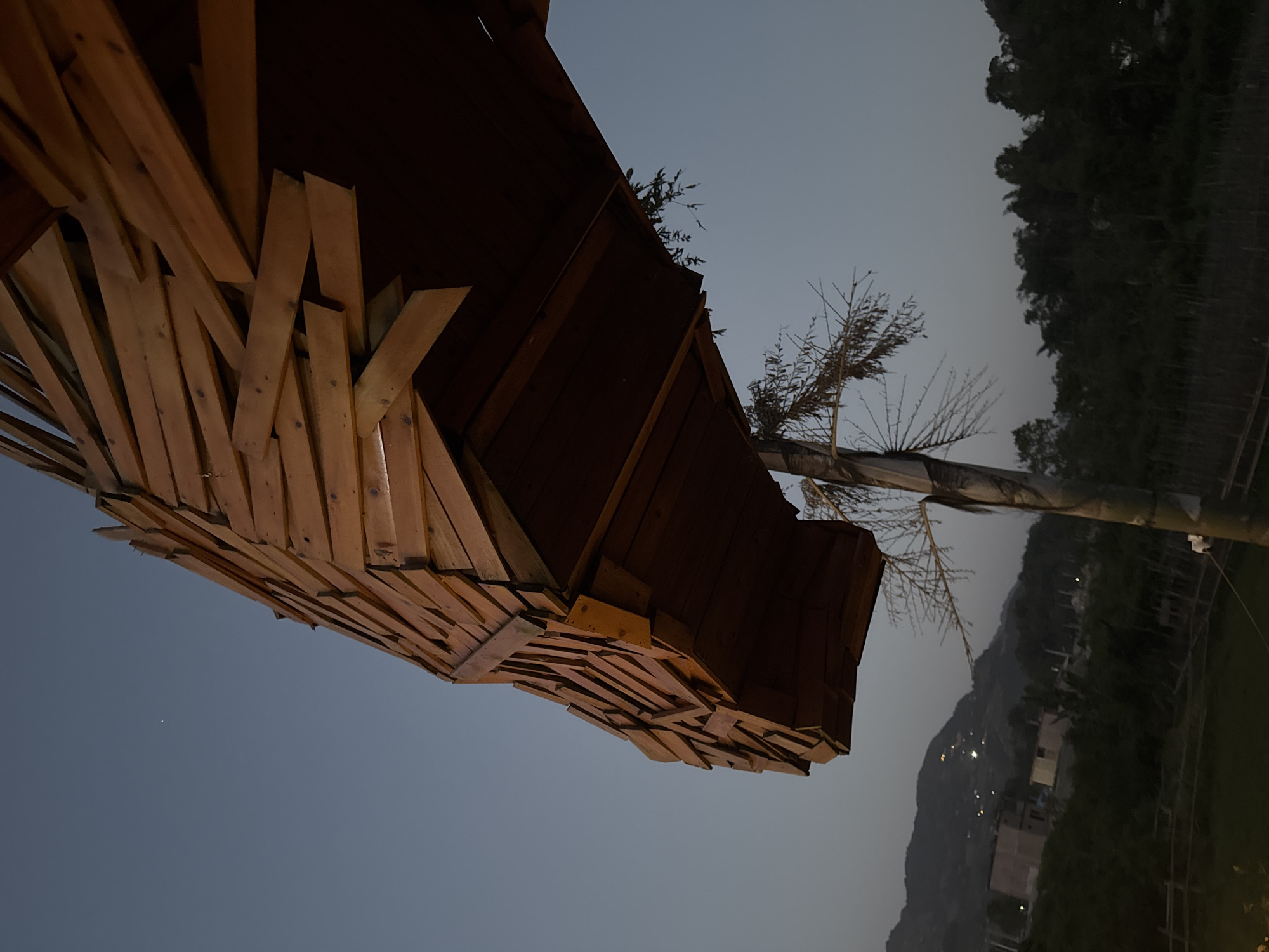The image captures a scene in Bắc Hà, Vietnam, at dusk or twilight. The foreground is dominated by a distinctive wooden structure, possibly a pavilion or architectural feature, with a complex design using numerous planks and beams. The color of the wood suggests it may be varnished or treated. Beyond the structure, a slender, bare tree stands against the evening sky. Further back, there's a silhouette of a mountain range and the presence of sparse buildings or lights scattered on the slopes, implying a residential area. The sky is a muted gradient of blue and gray, indicative of the fading daylight. The overall environment appears rural and mountainous, typical of the Bắc Hà region.