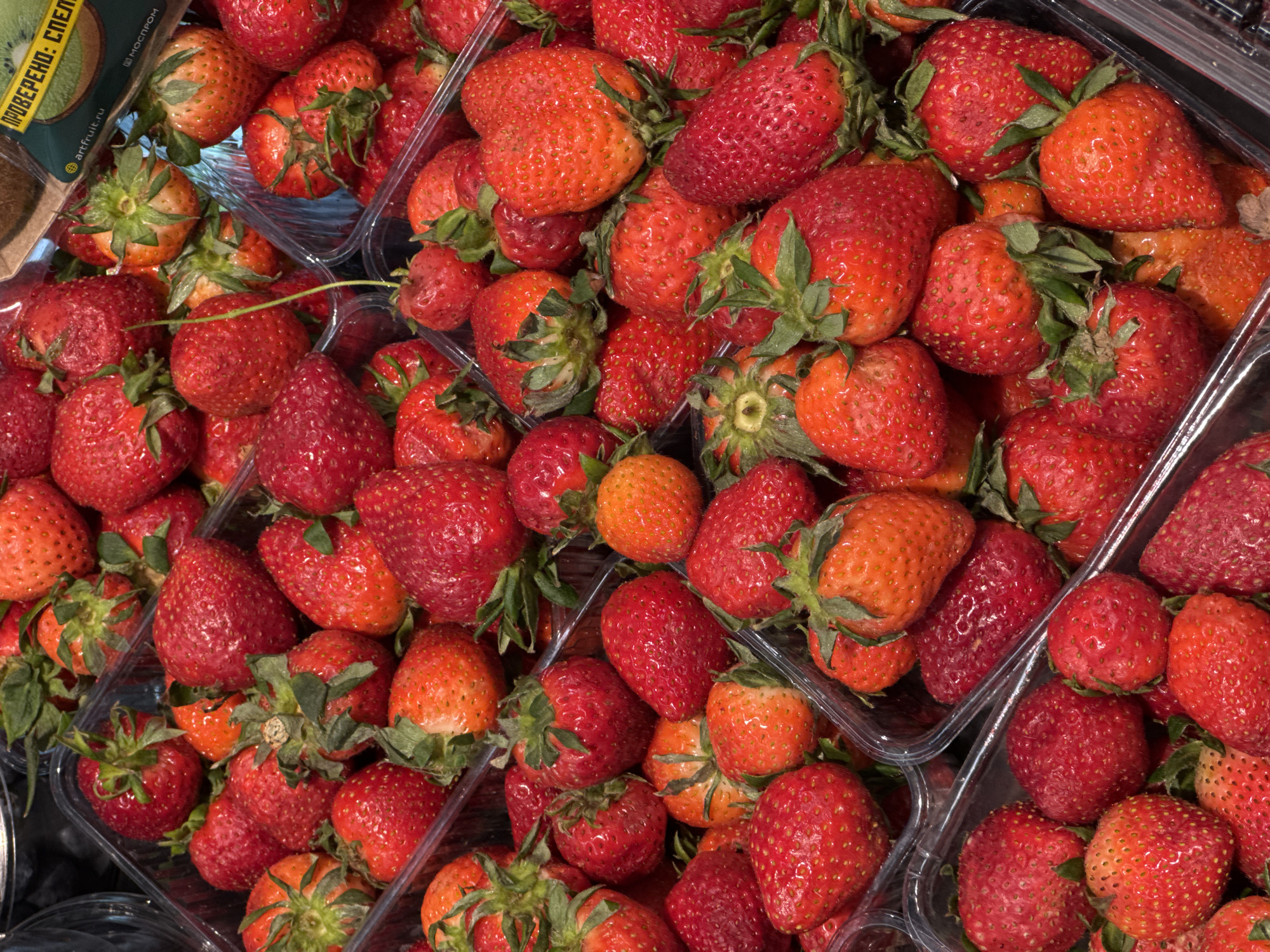 The image shows a close-up, overhead shot of several plastic containers filled with fresh strawberries. The strawberries vary in color from orange-red to deep red. Green leafy stems are visible on most of the strawberries. A corner of a cardboard box labeled in a Cyrillic language is visible in the upper left corner of the image. The location is unknown.