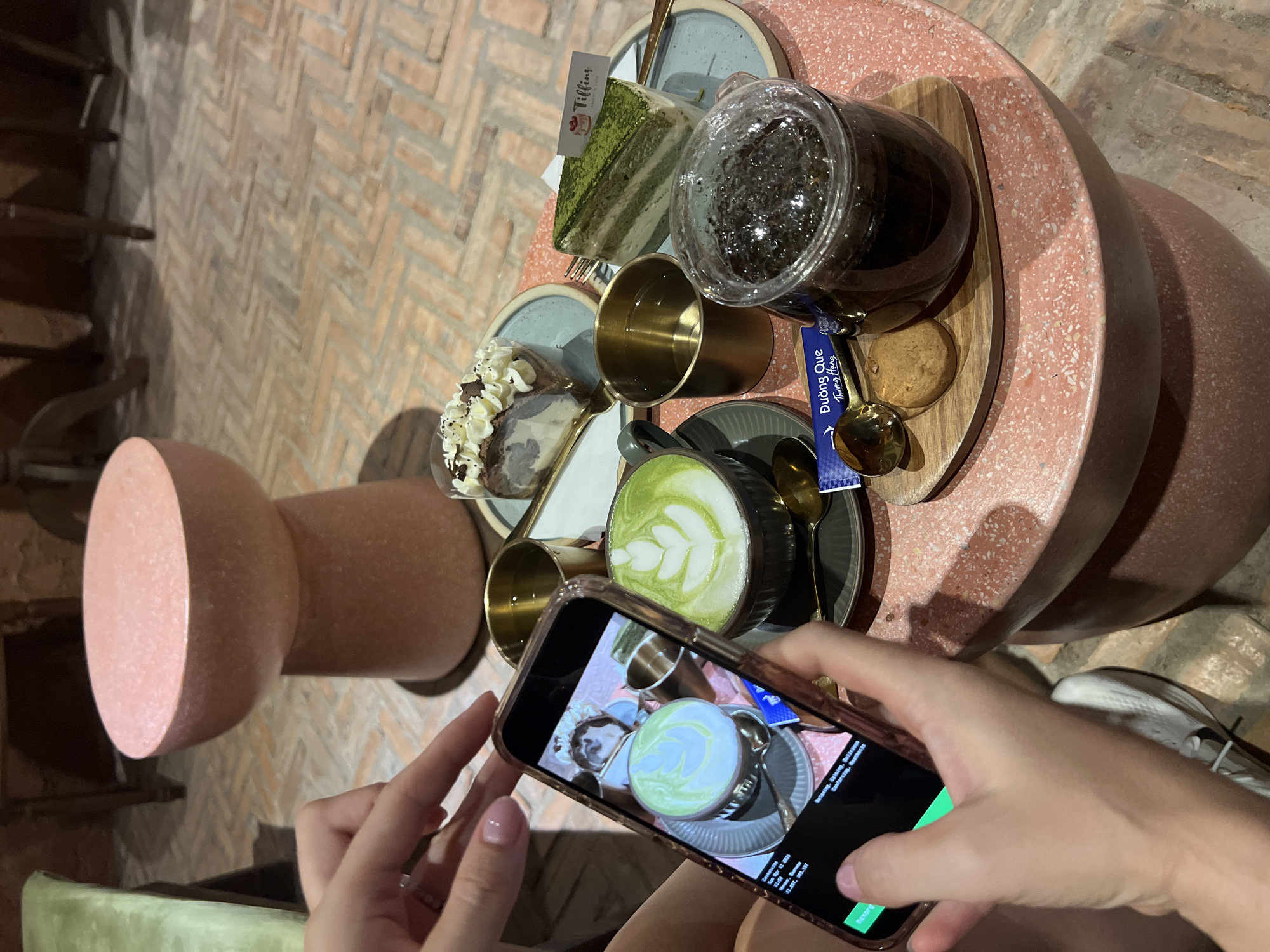 Captured from an elevated perspective, the image showcases a person using a smartphone to photograph a tabletop spread of refreshments in Bắc Nha Trang, Vietnam. The tabletop arrangement consists of a slice of green matcha cake with a "Tiffins" tag, a slice of chocolate cake, two gold-colored cups, a dark beverage in a clear jar, and a matcha latte. A wooden tray supports these items. The person's hands hold the smartphone, which displays a preview of the shot, while the legs of the person are visible, showing their attire and white sneakers. The table features a light-red, speckled surface, and the background reveals a terracotta-colored brick floor. Other elements include a section of a chair and the cylindrical table base. The atmosphere is casual and suggests an act of sharing visual content of the experience.